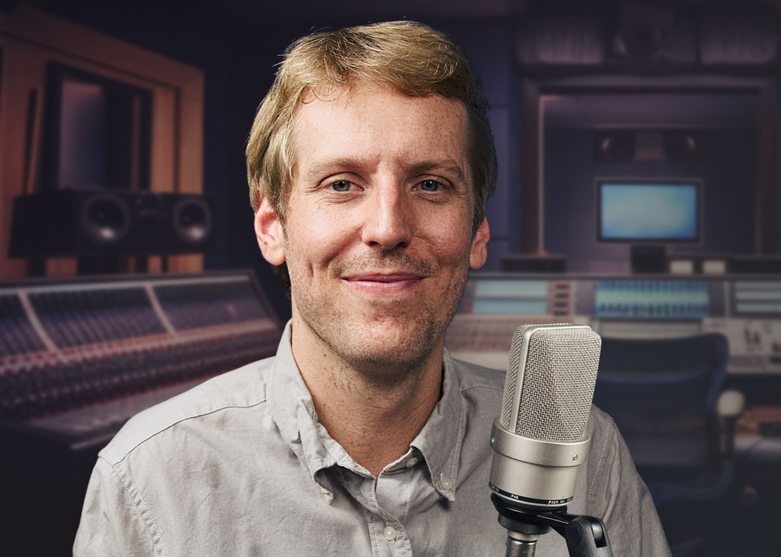 A man with short blonde hair smiling, standing in a recording studio with a large microphone in front of him, and sound mixing equipment and monitors in the background.
