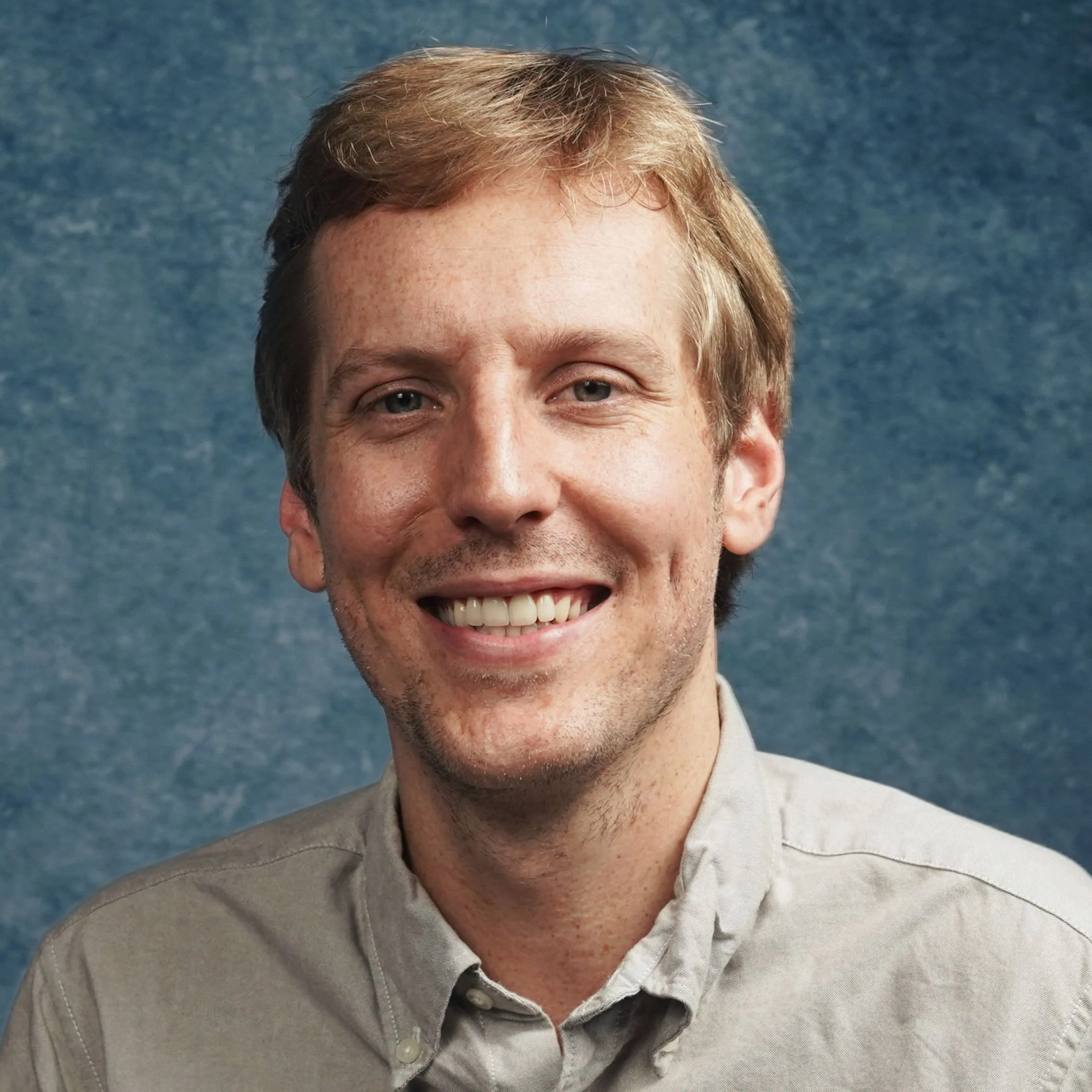 Headshot of a smiling man with light brown hair and blue eyes, wearing a light-colored button-up shirt, against a textured blue background.