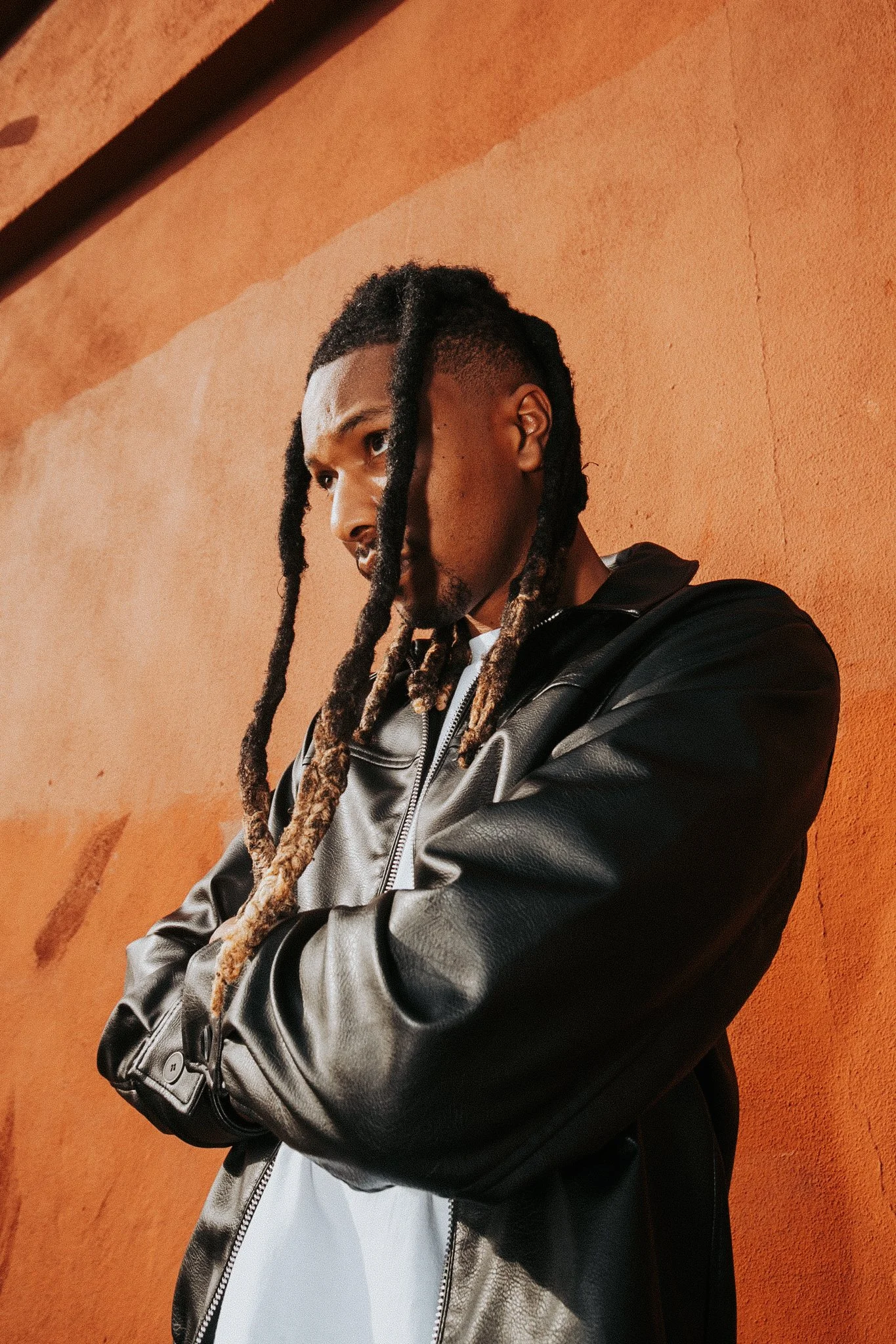 A person with long dreadlocks and a beard standing against an orange wall, wearing a brown and black leather jacket over a white shirt.