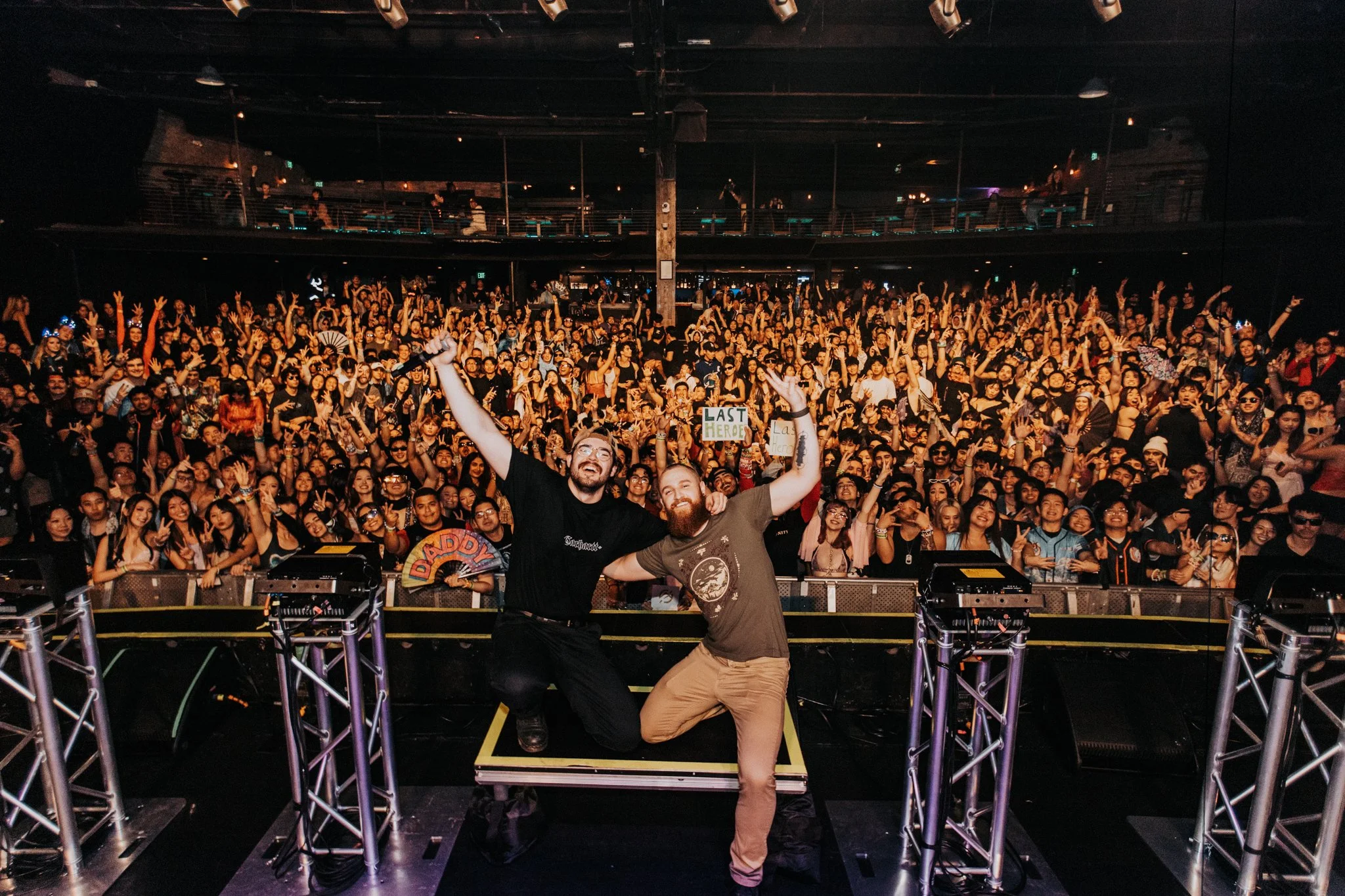 Two men pose smiling and making peace signs at a large concert audience, who are cheering and waving their hands in a dark indoor venue.