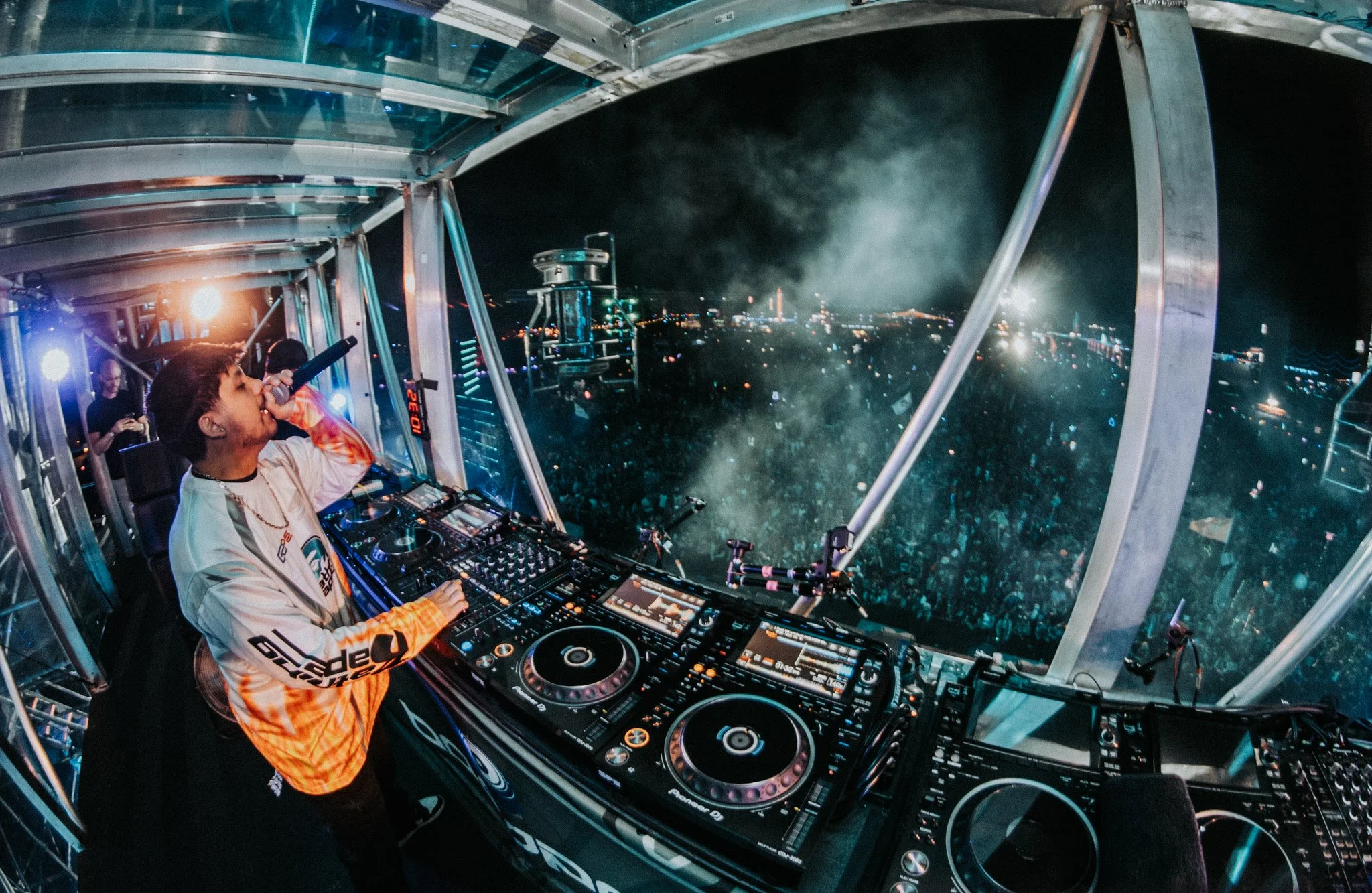 A DJ is performing at a night event on a glass platform high above a cityscape. The DJ is using a Pioneer DJ setup, wearing a white and orange jersey, with city lights and smoke in the background.