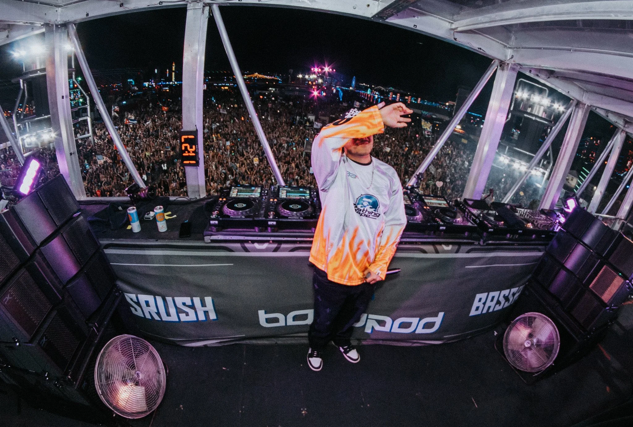 A DJ standing in front of a large crowd at a nighttime outdoor music festival, with city lights in the background.
