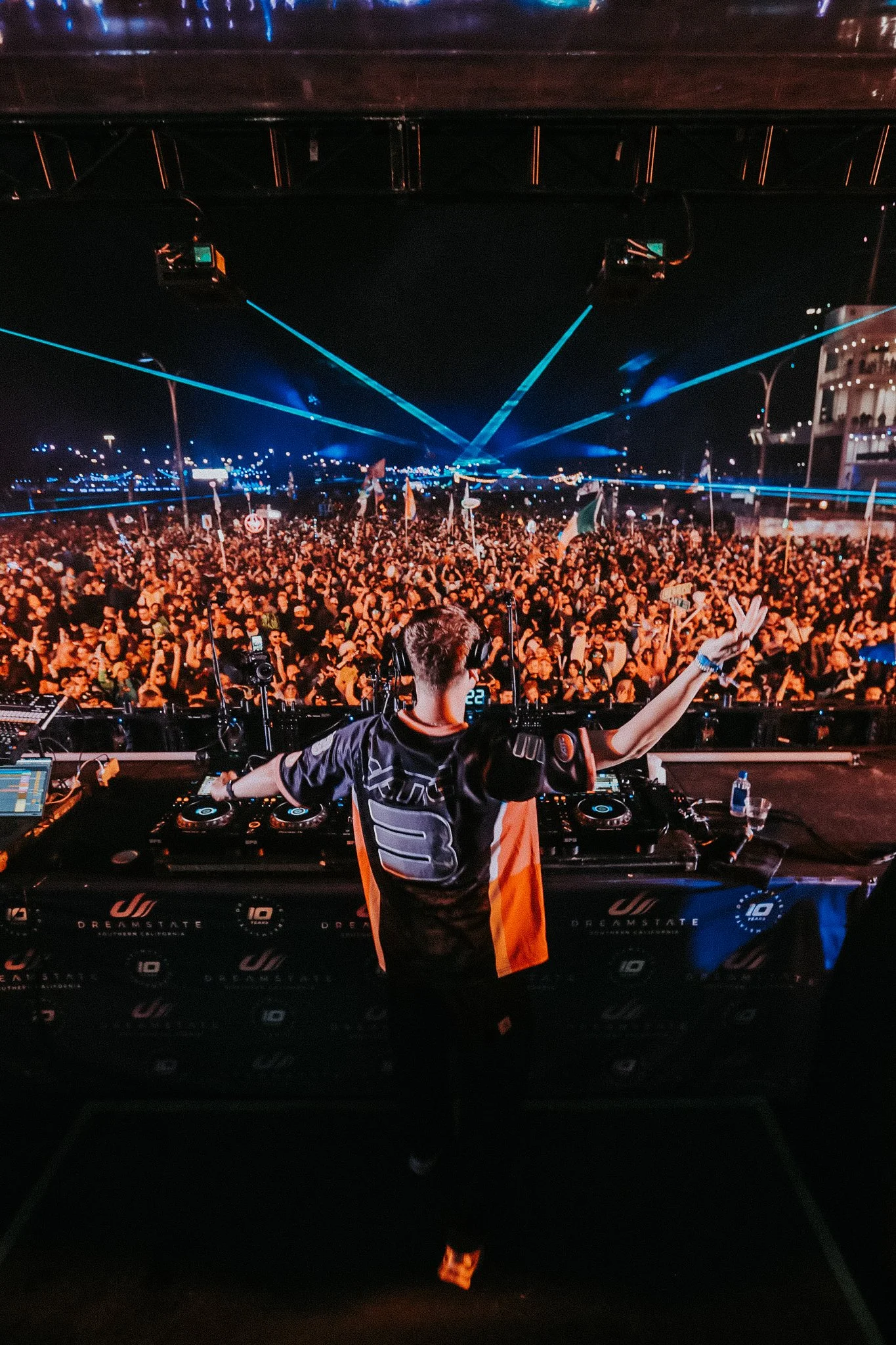 A DJ performing on stage at night with a large crowd and blue laser lights.