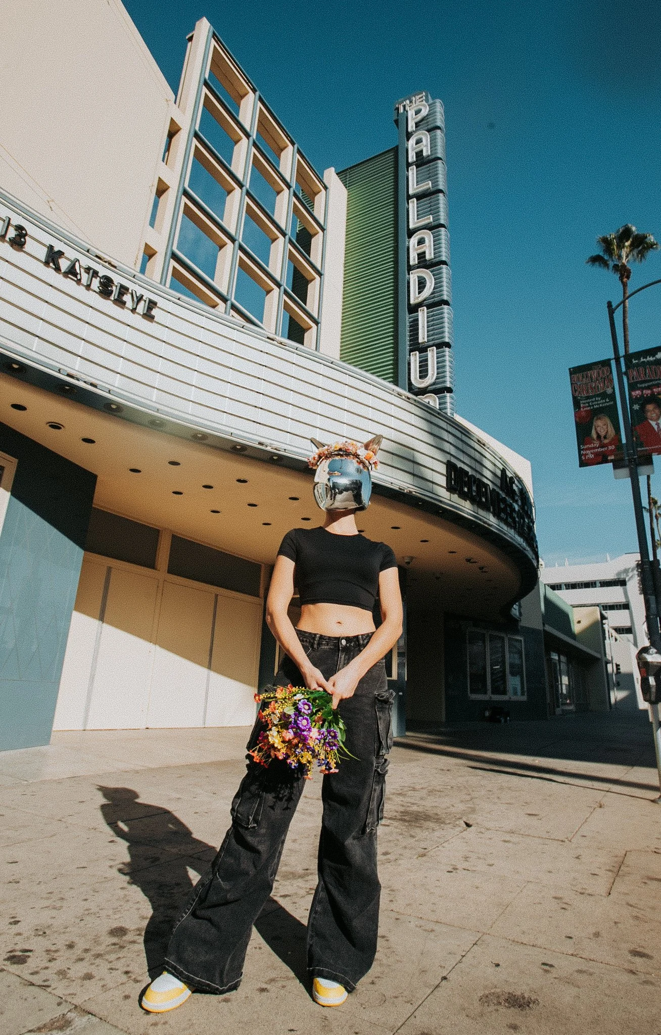 Person standing outside the Paley Theater holding a bouquet of flowers, wearing a reflective helmet, black crop top, wide black pants, and yellow-and-white sneakers, with the theater marquee and sign visible in the background.
