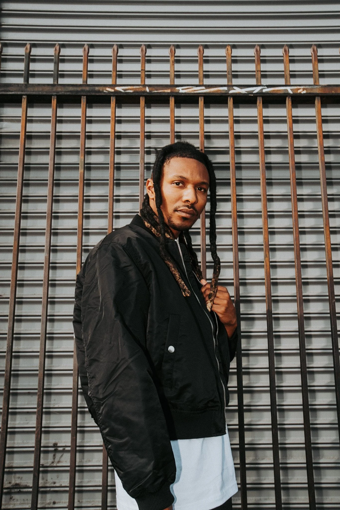A young man with long dreadlocks wearing a black jacket and white shirt standing in front of a metal gate with horizontal and vertical bars.