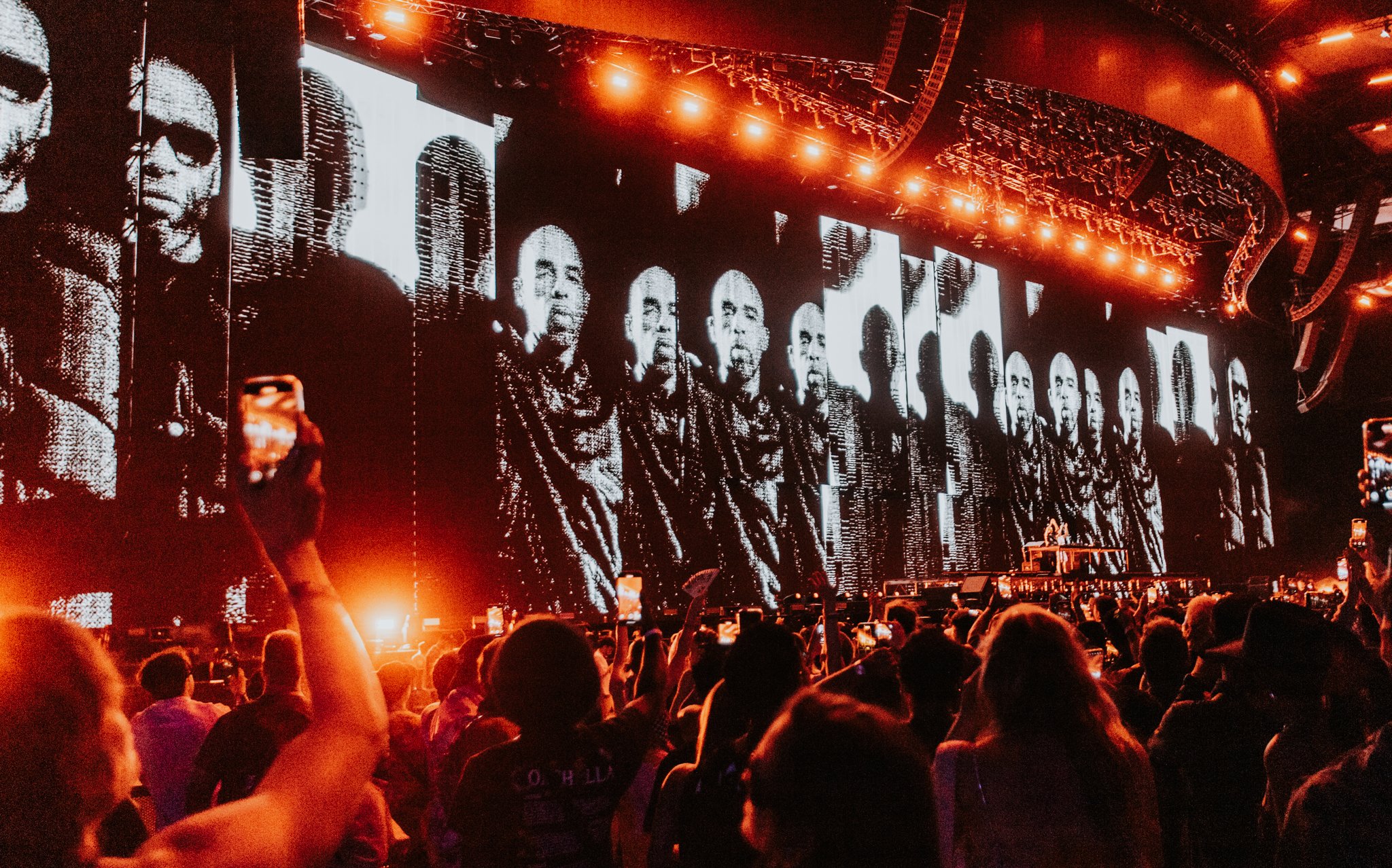 A large crowd at a concert with multiple big screens showing black and white images of a man wearing sunglasses, with orange stage lighting overhead.