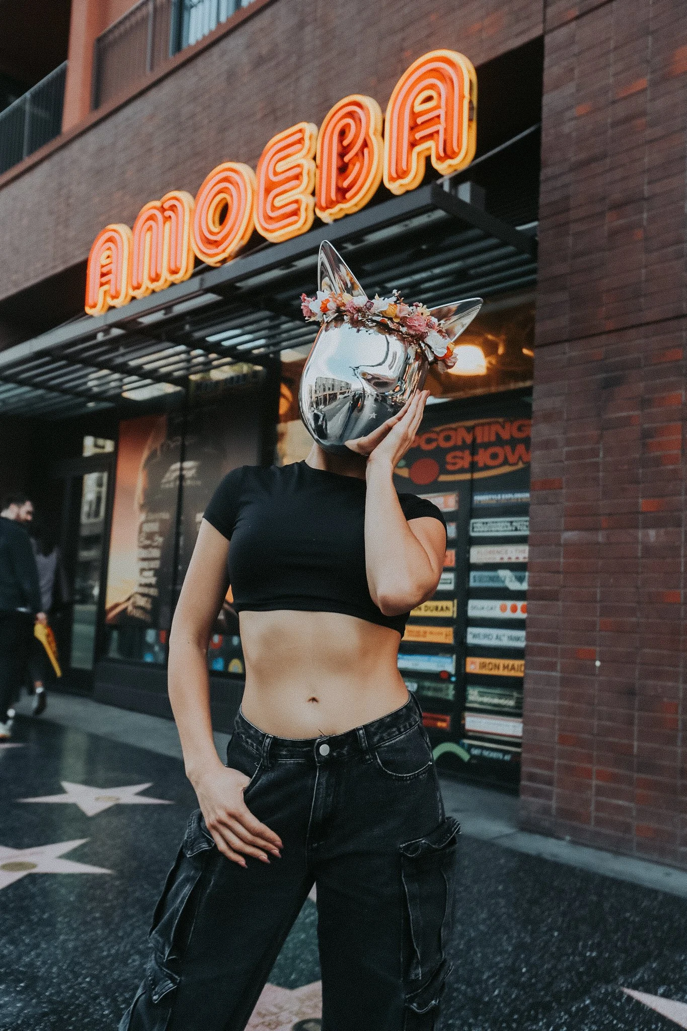 Person with a metallic dog-shaped mask with a flower crown, wearing a black crop top and black leather pants, standing in front of a building with a neon sign that says 'AMOEBA'.