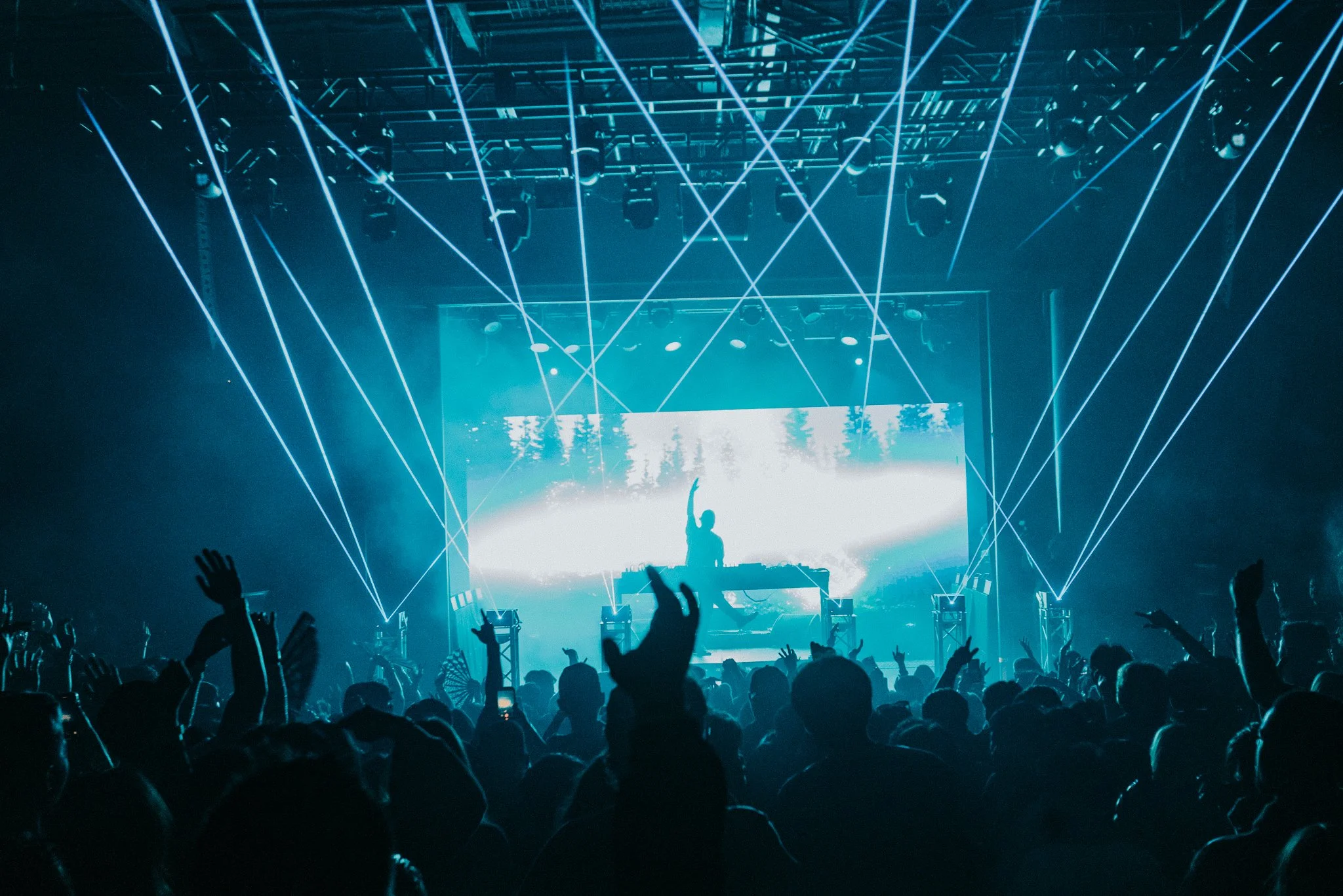A concert scene with a DJ on stage, large crowd with hands raised, blue laser lights crisscrossing above, and a large screen displaying a scenic background.