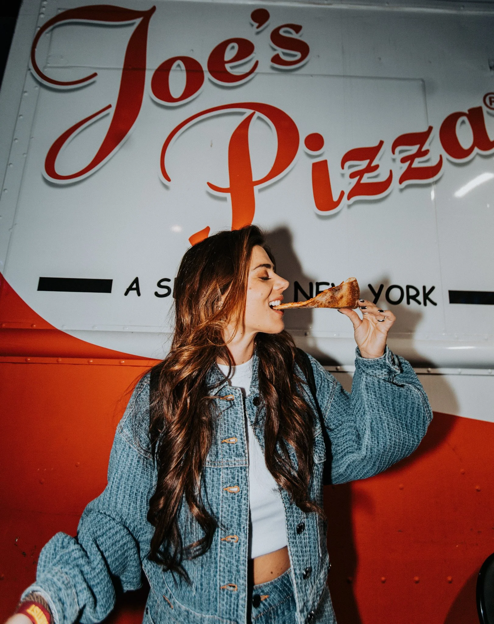 A woman with long, wavy brown hair wearing a denim jacket and white top, holding a slice of pizza near her mouth, standing in front of a sign that reads 'Joe's Pizza.'