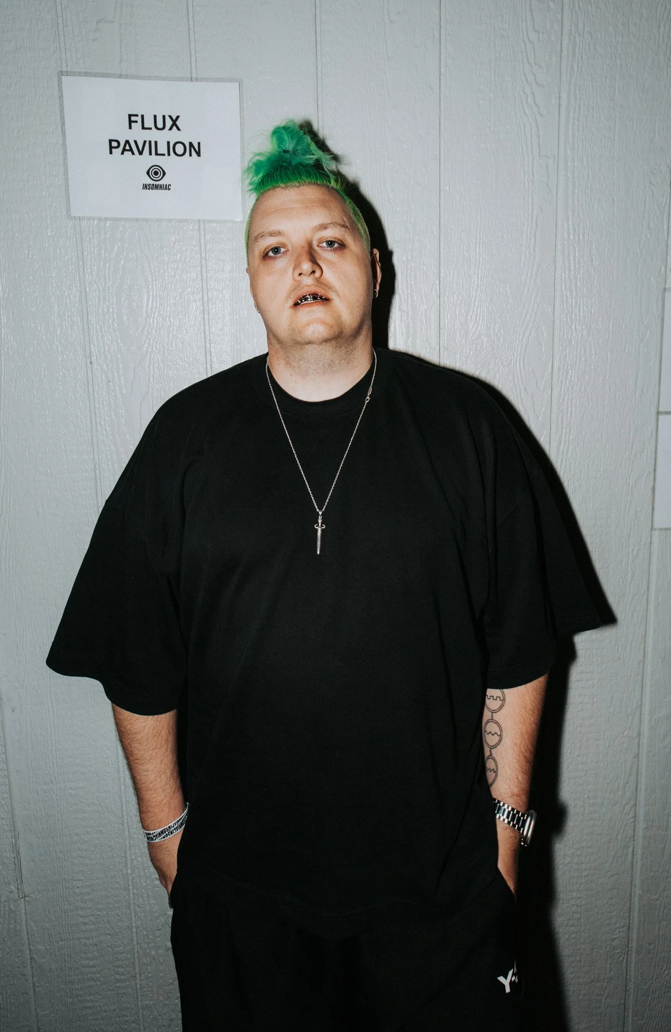 A young man with green hair styled in a top knot, wearing a black T-shirt and a silver necklace, standing against a gray wall with a sign that reads 'Flux Pavilion' and 'Insomniac.'
