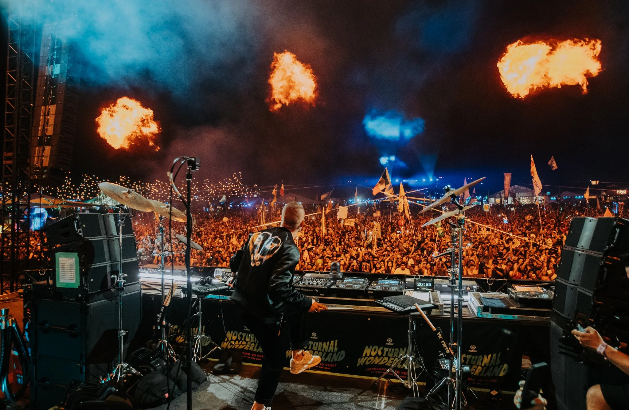 A DJ performs on stage at a large outdoor concert or music festival at night, with a crowd of people in front and decorative flames in the sky.