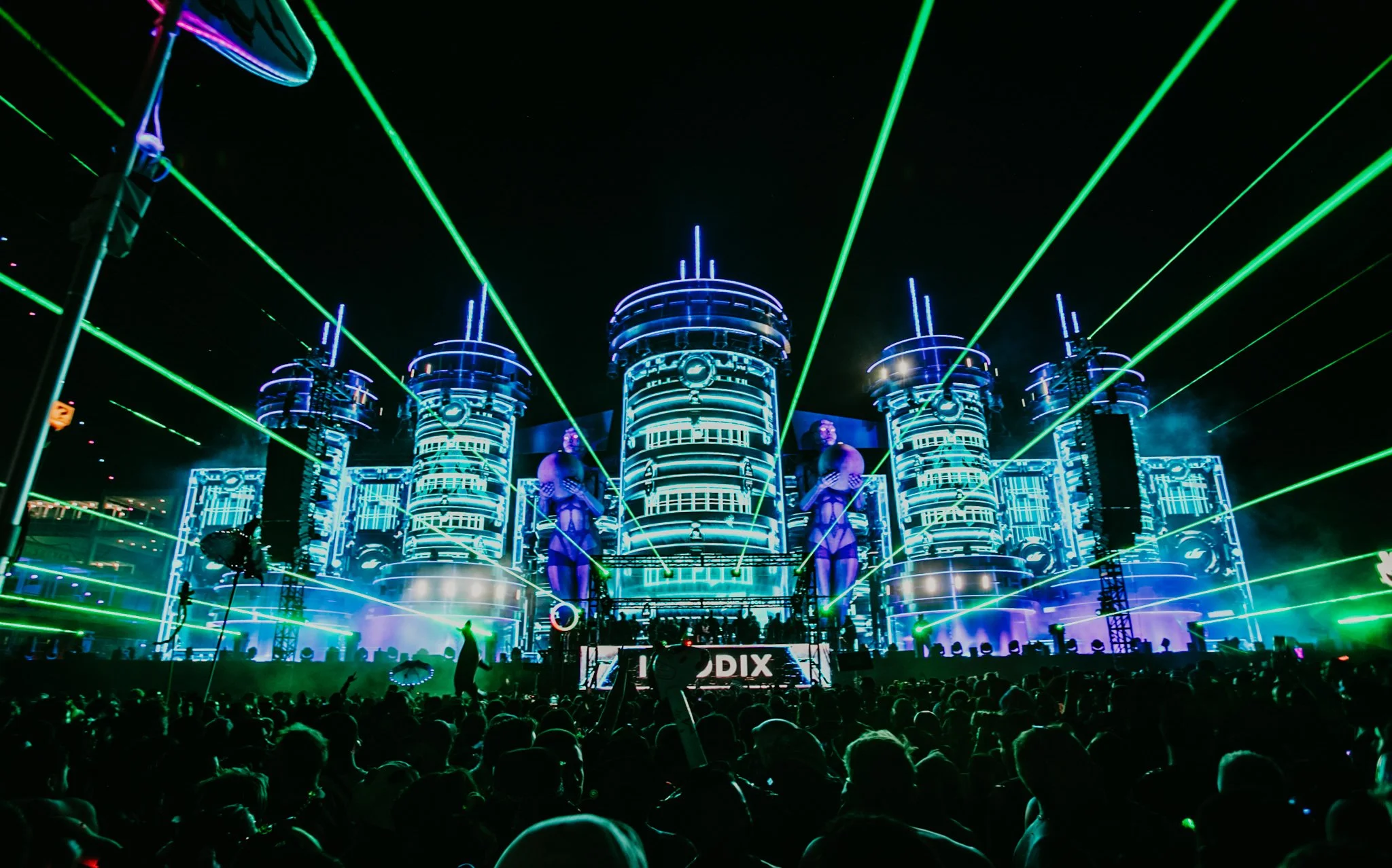 A daytime scene at an outdoor music festival with a large stage featuring futuristic, cylindrical LED structures and detailed light displays. The stage is illuminated with green laser beams, and a crowd of festival-goers is visible in the foreground,