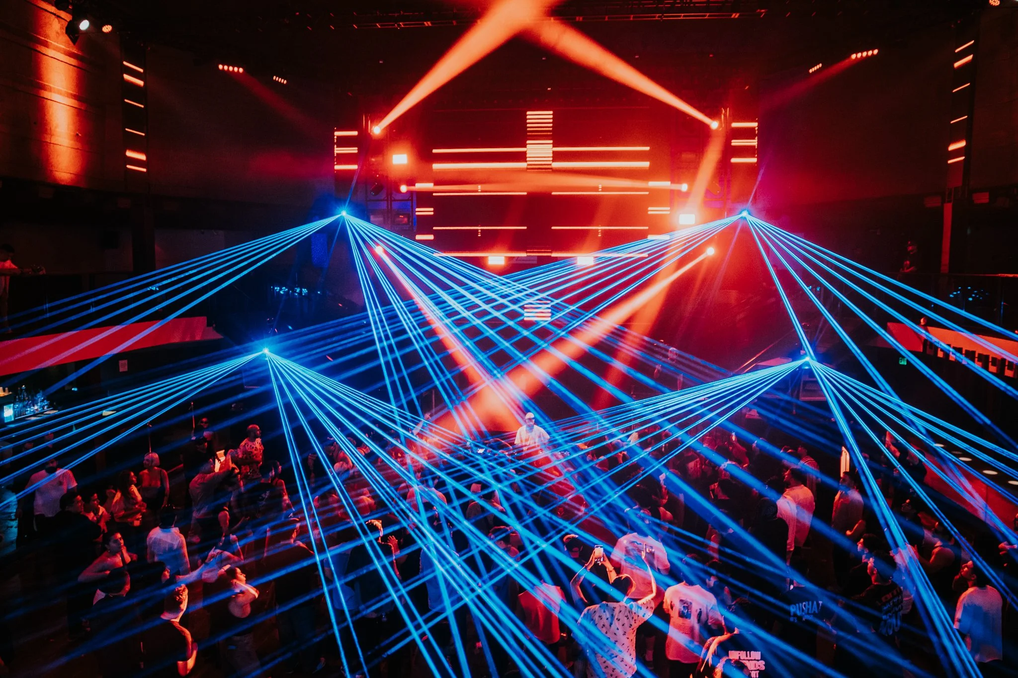Dance club scene with blue laser lights crossing over an audience in front of a stage with red lighting and digital screens.