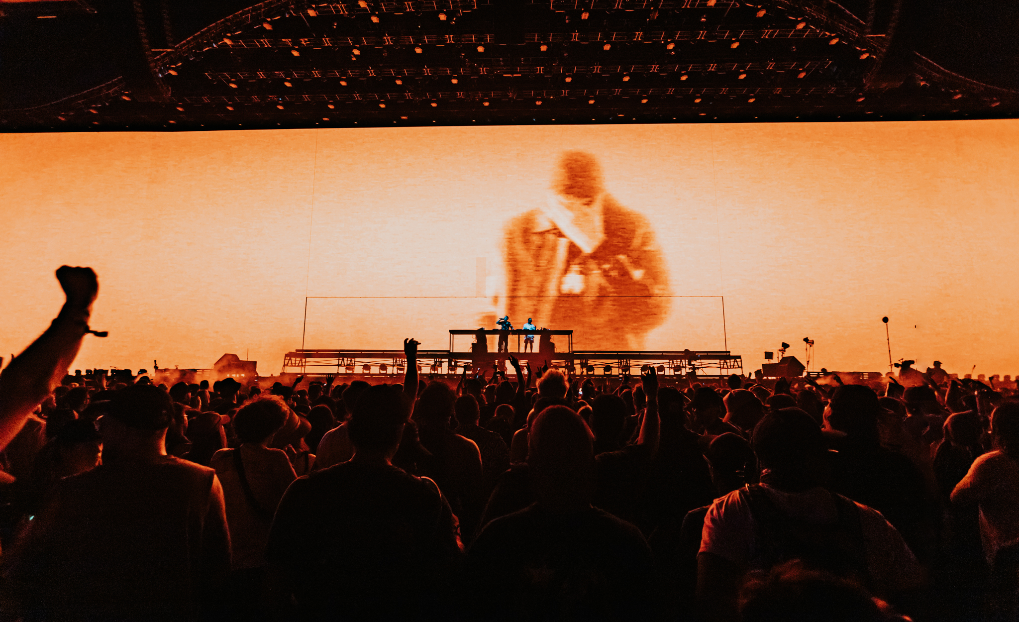 Crowd of people at a concert with a large orange screen displaying a blurry image of a person.