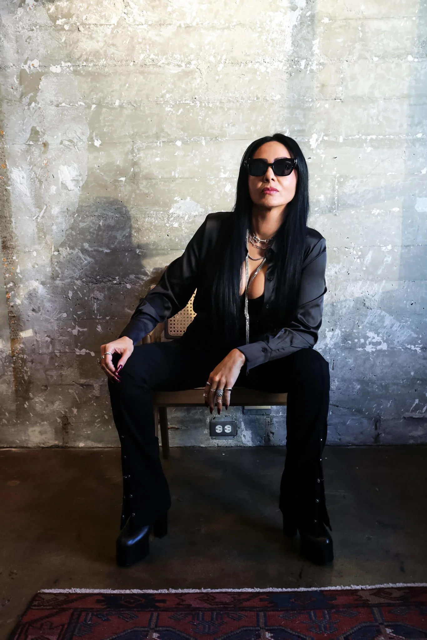 A woman with long black hair wearing black sunglasses, a black leather jacket, and black pants, sitting on a chair against a weathered concrete wall, with a power outlet visible on the wall below her. She has a serious expression and is accessorized 