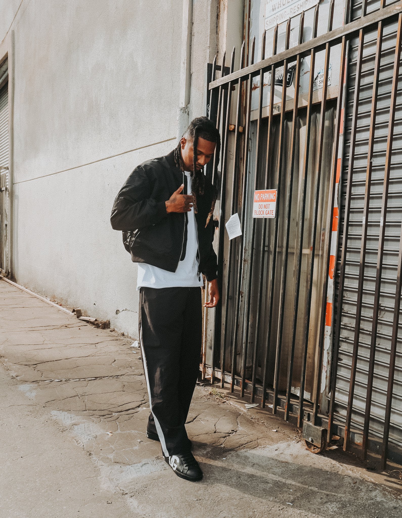 A young man with dreadlocks wearing a black jacket and black and white pants stands on a cracked sidewalk near a metal gate, looking down at the ground.