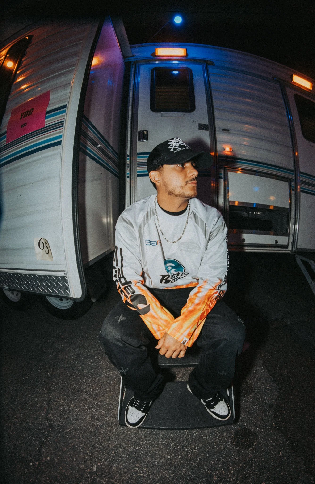 A young man wearing a black cap, a racing jersey, and black pants, sitting on a stool in front of an RV at night, with blue and orange lights reflecting off the RV.