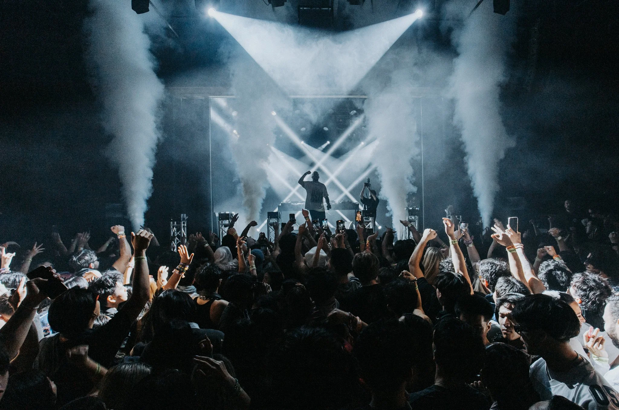 A large crowd at a music concert with the stage illuminated by bright spotlights and smoke effects. The audience is cheering and raising their hands.