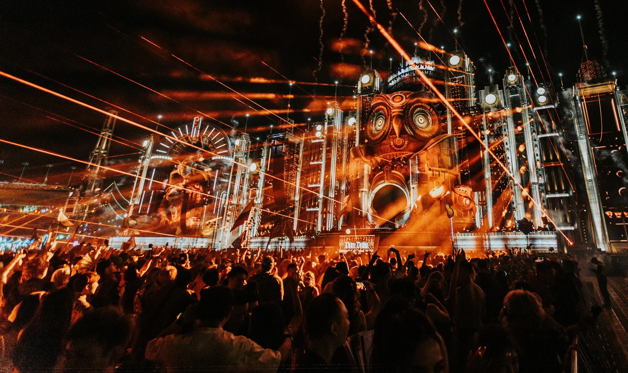 A large crowd at night at an electronic music festival with bright orange and white laser light shows and a stage decorated with an owl face.