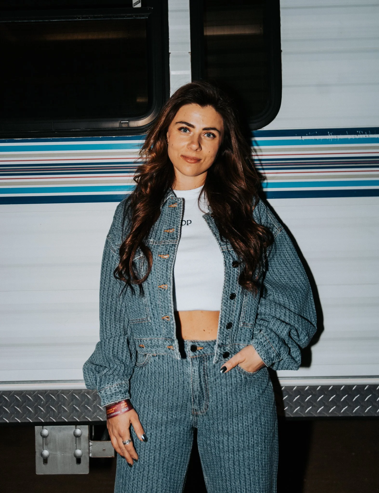 A woman with long brown hair and blue eyes, wearing a denim jacket and matching jeans, standing against a striped background. She is looking at the camera with a slight smile and has her right hand resting on her hip.