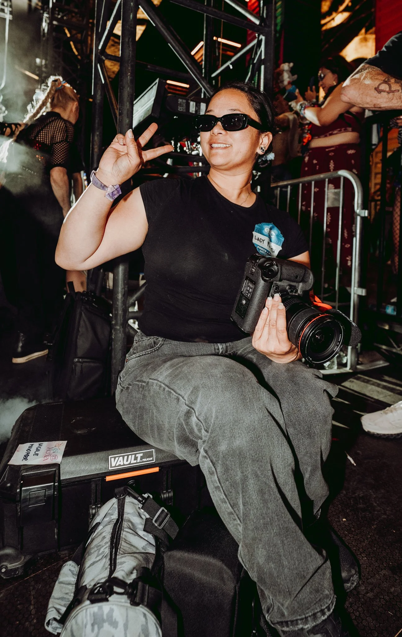 A woman with short dark hair, wearing black sunglasses and a black t-shirt, is sitting and smiling while making a peace sign with her right hand, holding a camera in her left hand. She has a tattoo on her right arm and is surrounded by other people and equipment in an indoor setting.