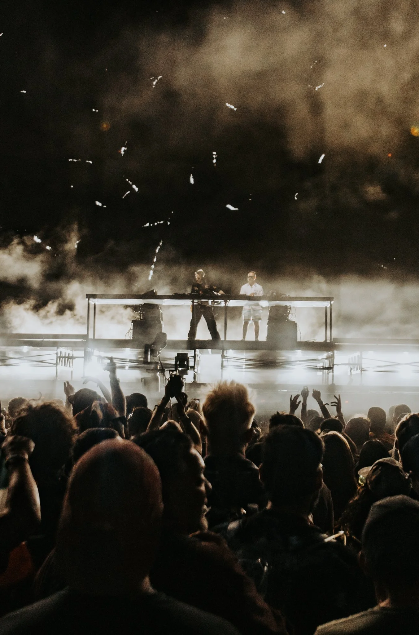 Crowd watching a concert with two DJs on stage, fog effects, and fireworks in the background at night.