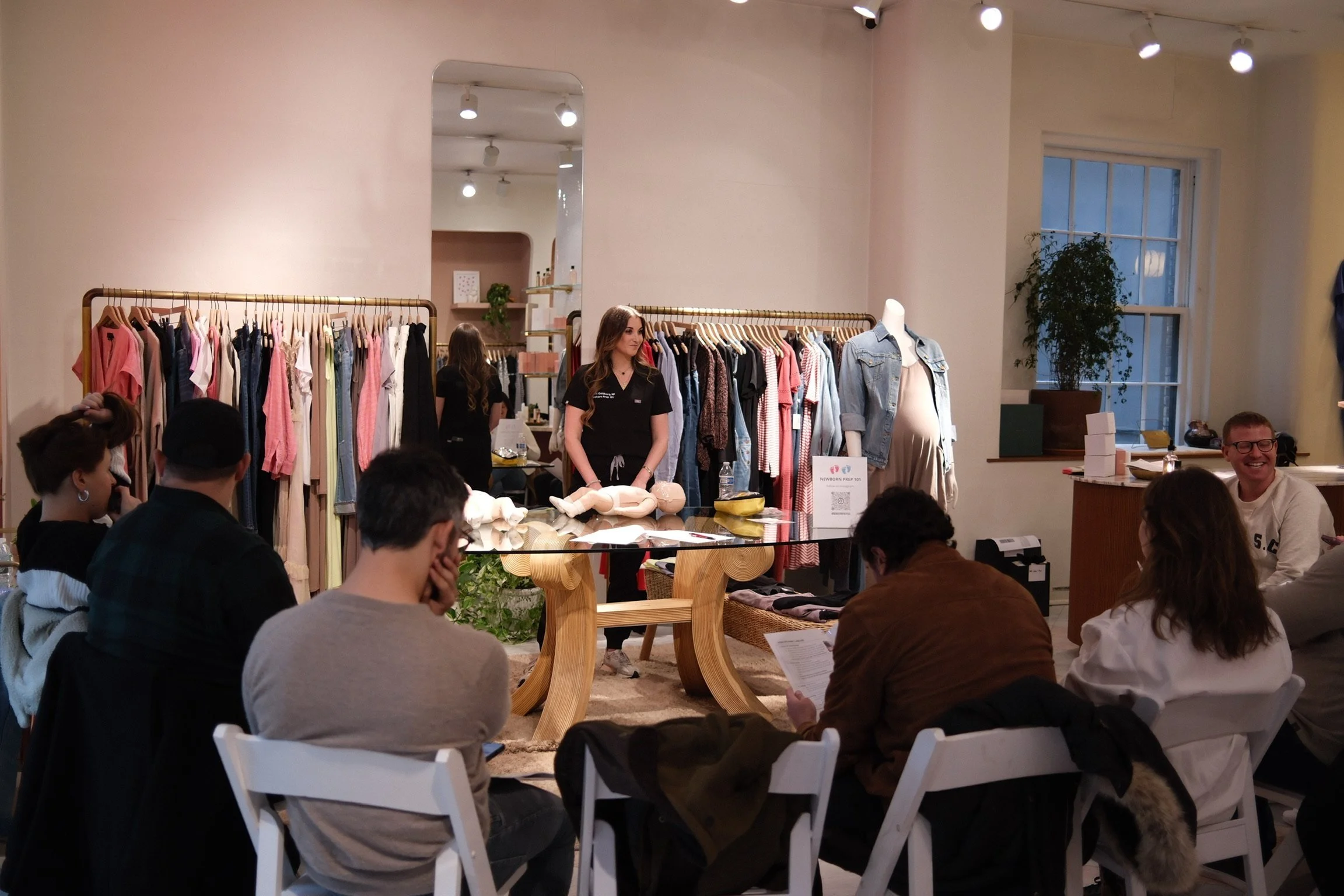 A woman presents a parenting class with a newborn doll demonstration in a boutique with clothing racks and mannequins, audience seated at white chairs, and smiling participants.