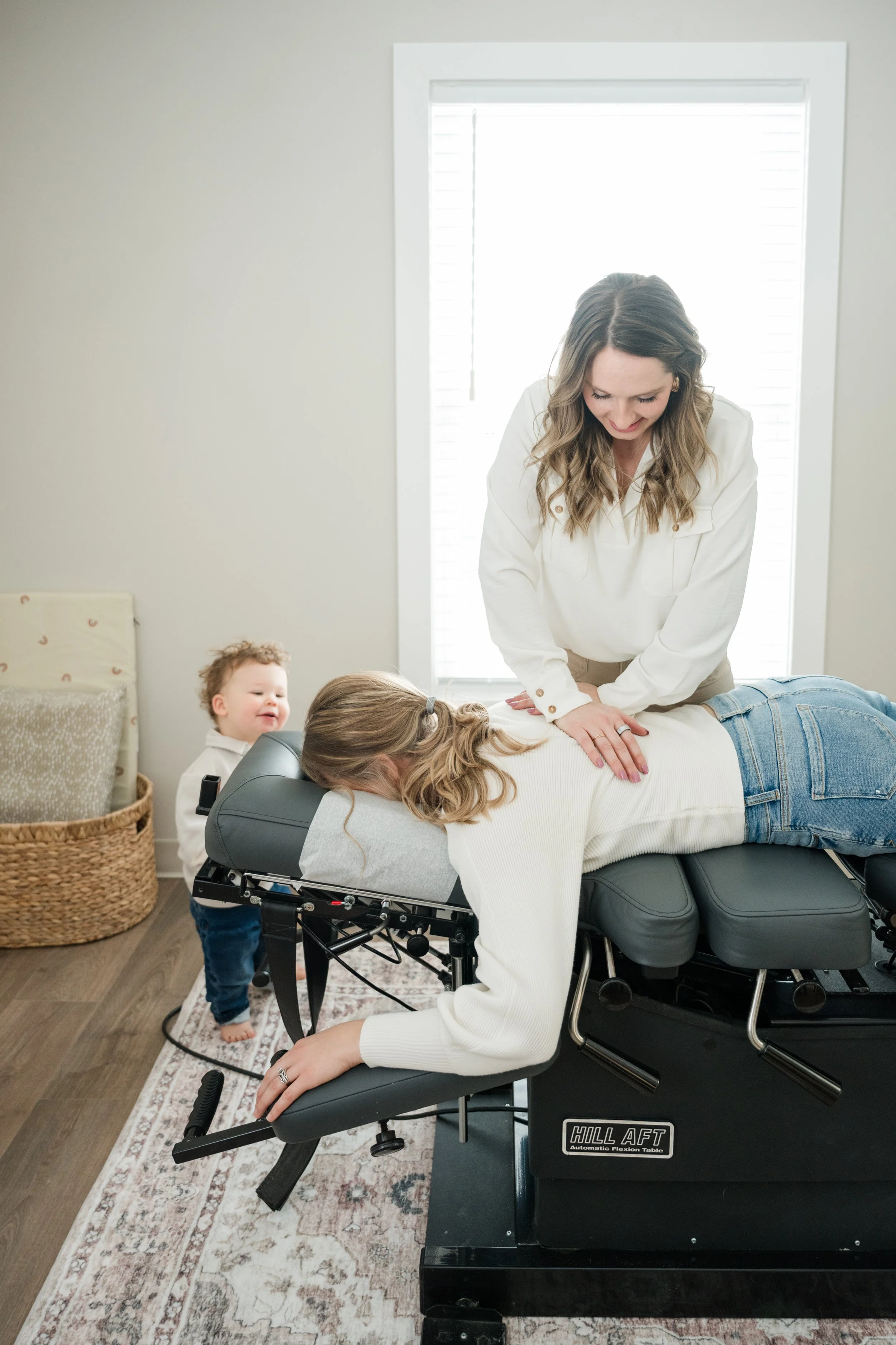 A woman receiving a chiropractic adjustment from a female chiropractor in a clinic while a young child watches and smiles in the background.