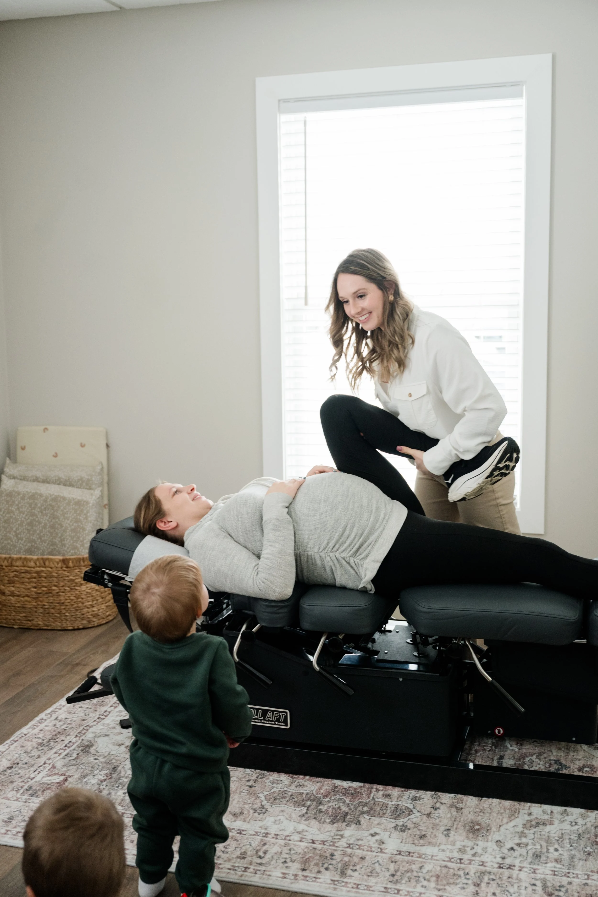 A woman lying on a chiropractic adjusting table with her eyes closed, a young woman standing next to her smiling, and two small children observing in a room with a window and a basket in the background.