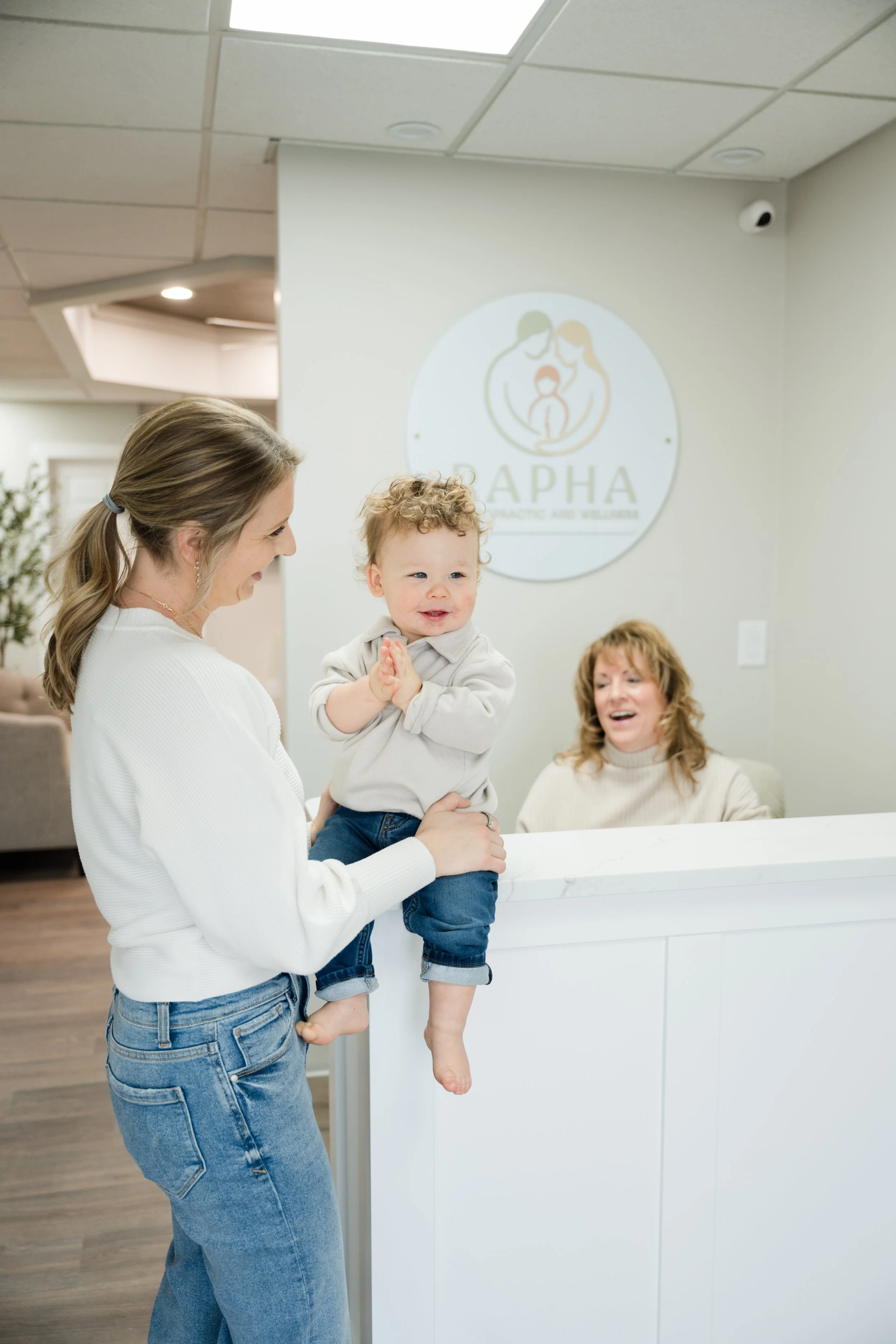 A woman is holding a young boy at a reception desk, smiling, while a woman behind the desk looks on and laughs. A logo for "Alpha Chiropractic and Wellness" is visible on the wall behind them.
