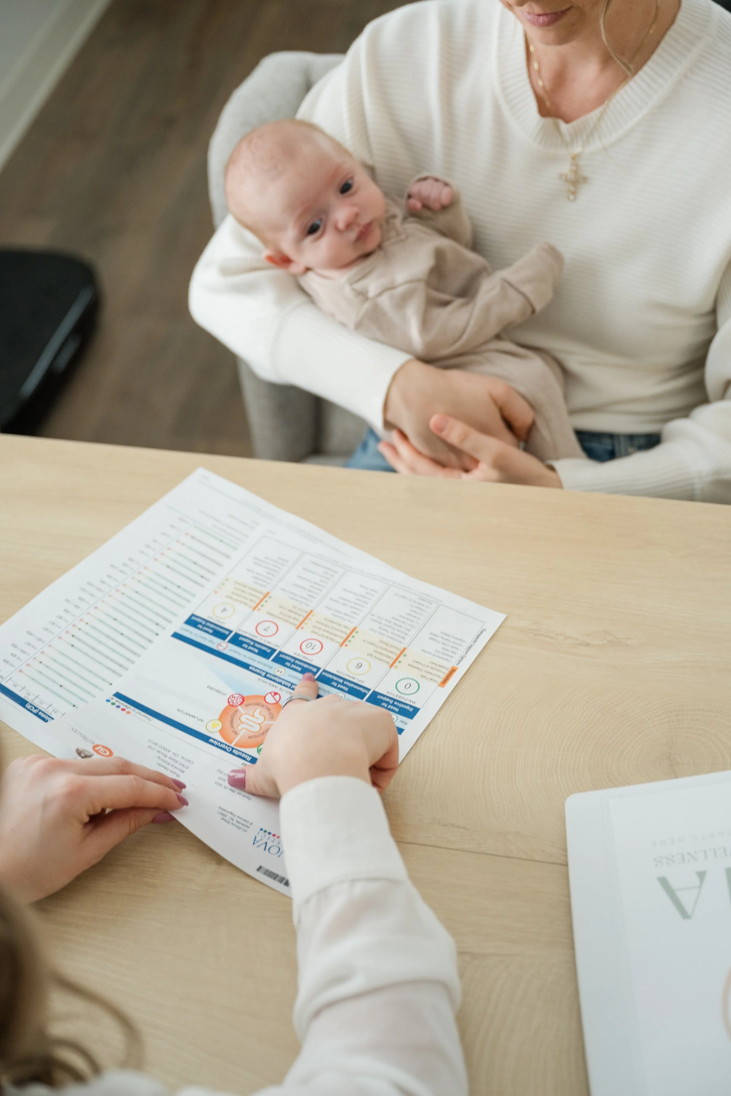 A woman holding a baby while a person points at a health test or vaccination card on a wooden table.
