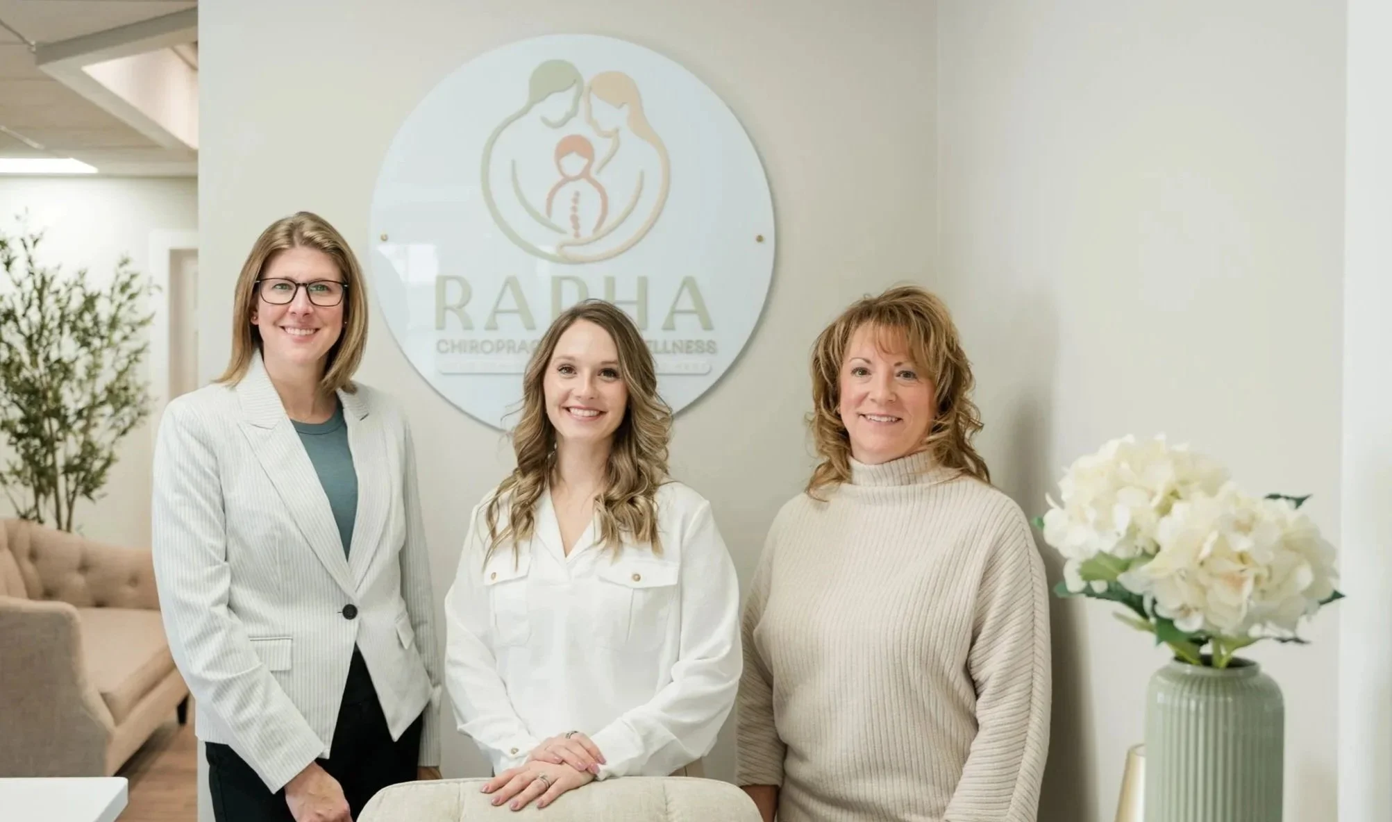 Three women standing together in an office reception area, smiling. A logo with a woman and child silhouette and the words "RADA Chiropractic Wellness" is on the wall behind them. There is a green vase with white flowers on a table to the right.
