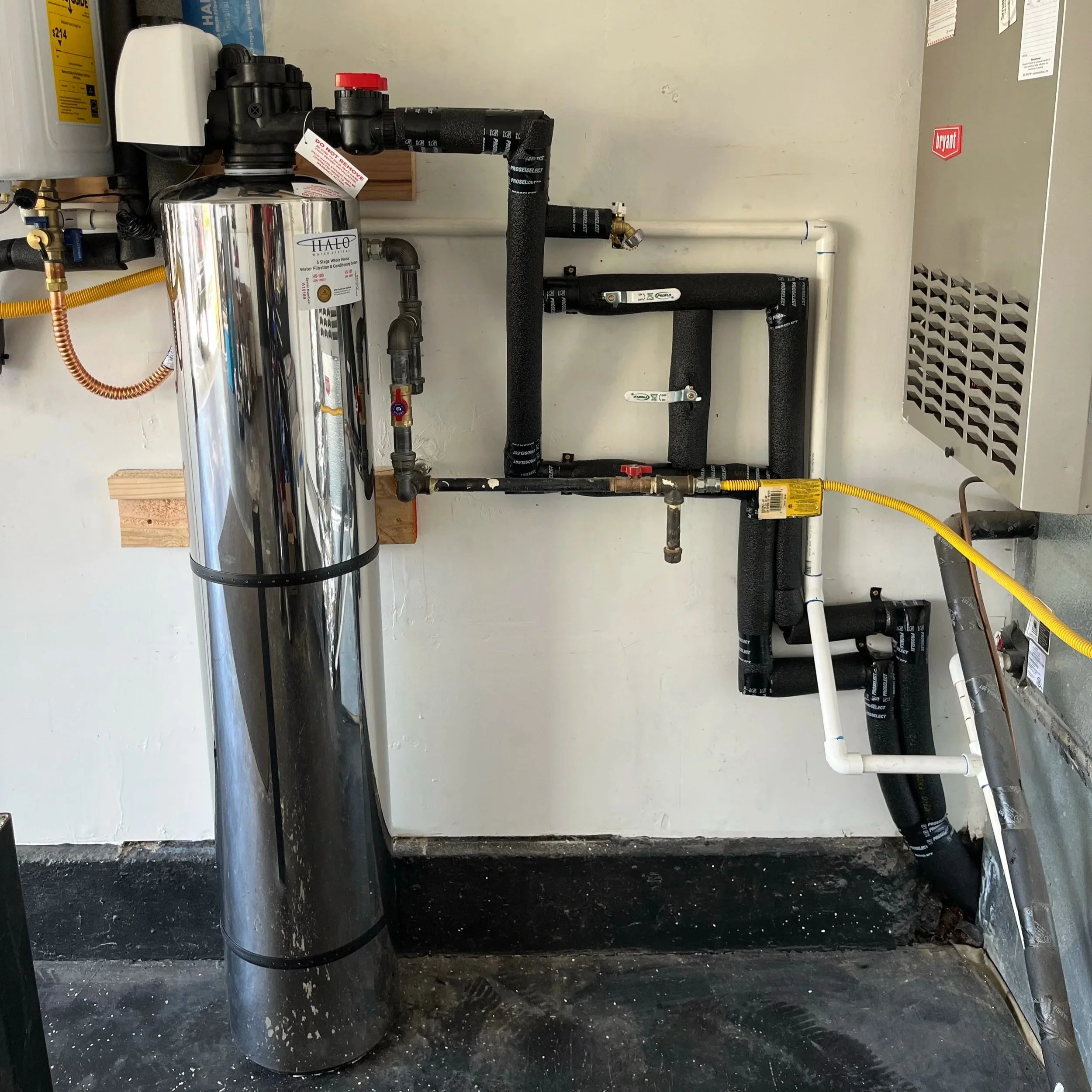 A utility room with a large, shiny, chrome water filter tank mounted on a black pipe system, connected to various black, white, and yellow pipes and valves. There is a gray HVAC unit on the right side of the wall and a wooden platform beneath the system. The floor is concrete with some dirt, and the background wall is plain painted white.