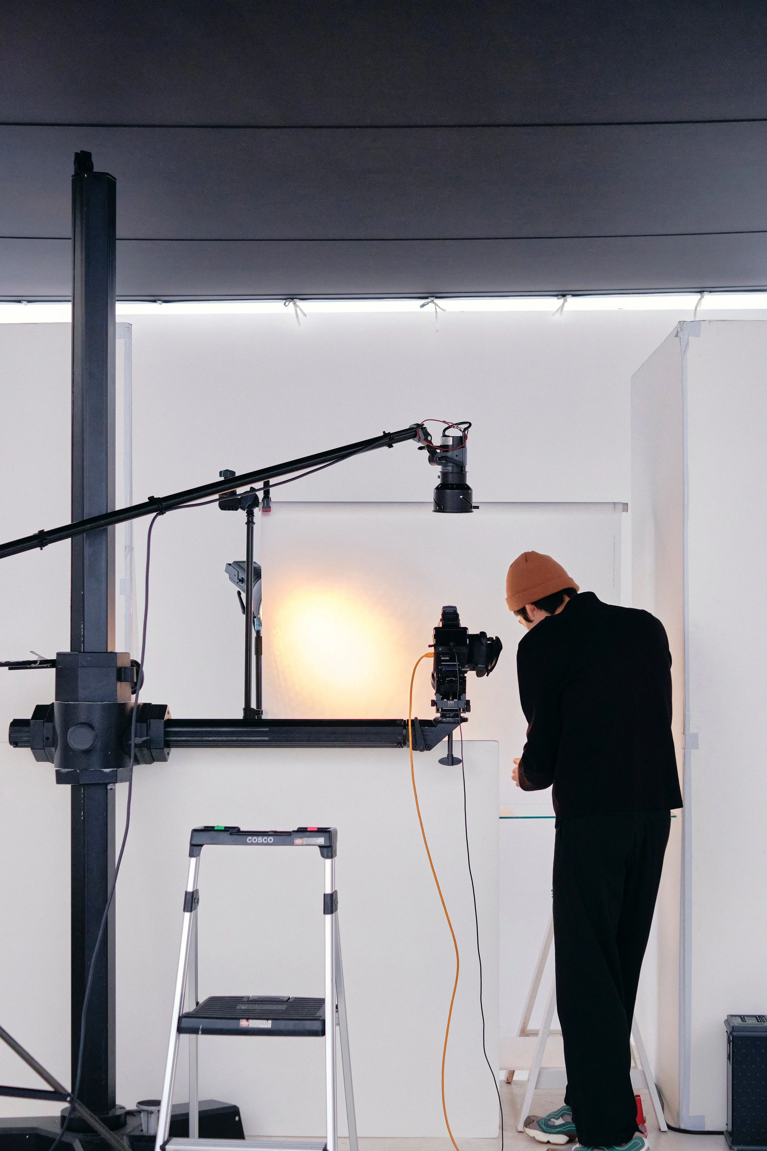 Person in black clothing and brown beanie adjusting equipment in photography studio with white backdrop, professional lighting and camera setup.