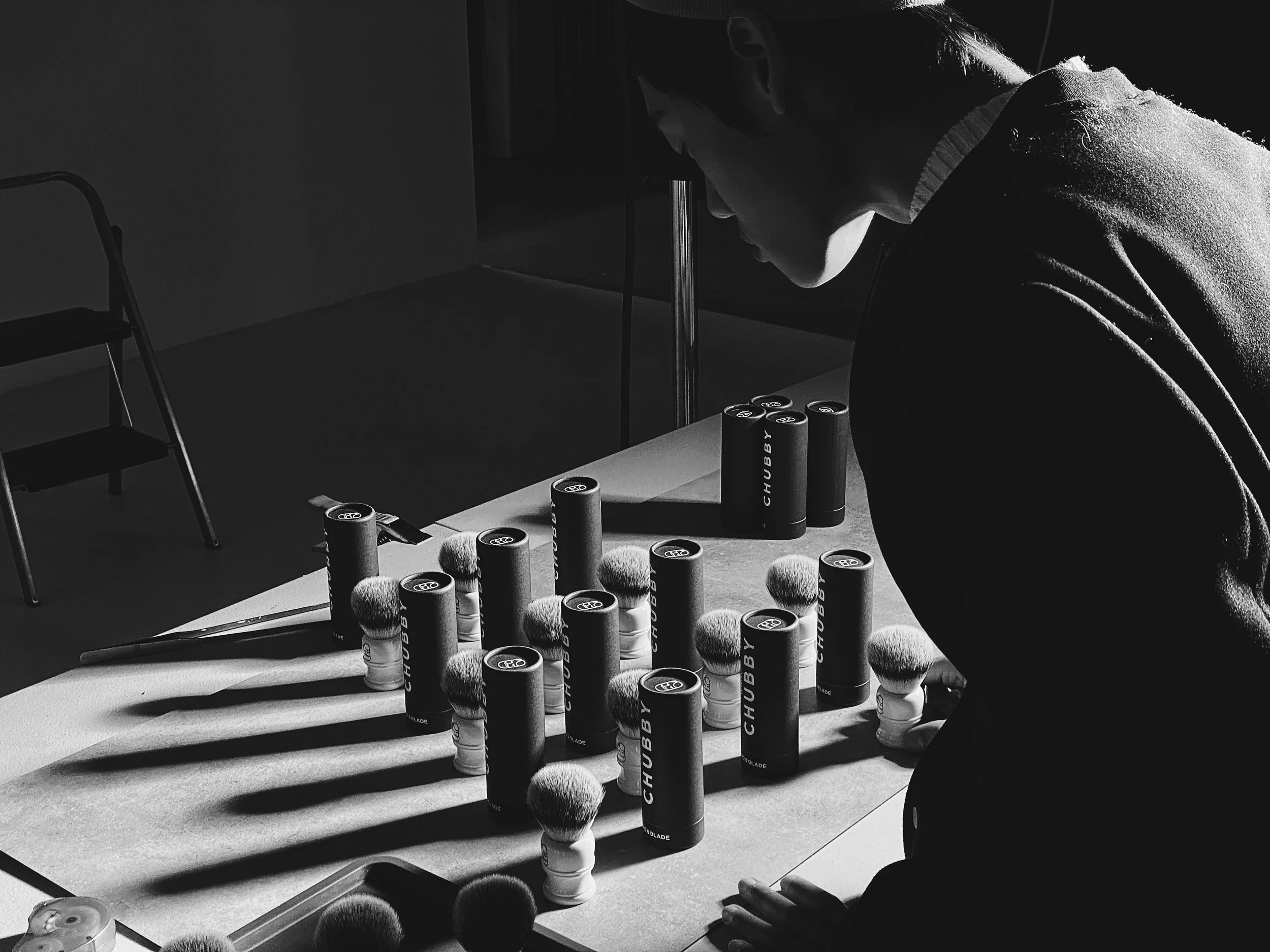 A person in profile leans over a table with shaving brushes and cans of shaving foam, with shadows cast from the brushes and cans.
