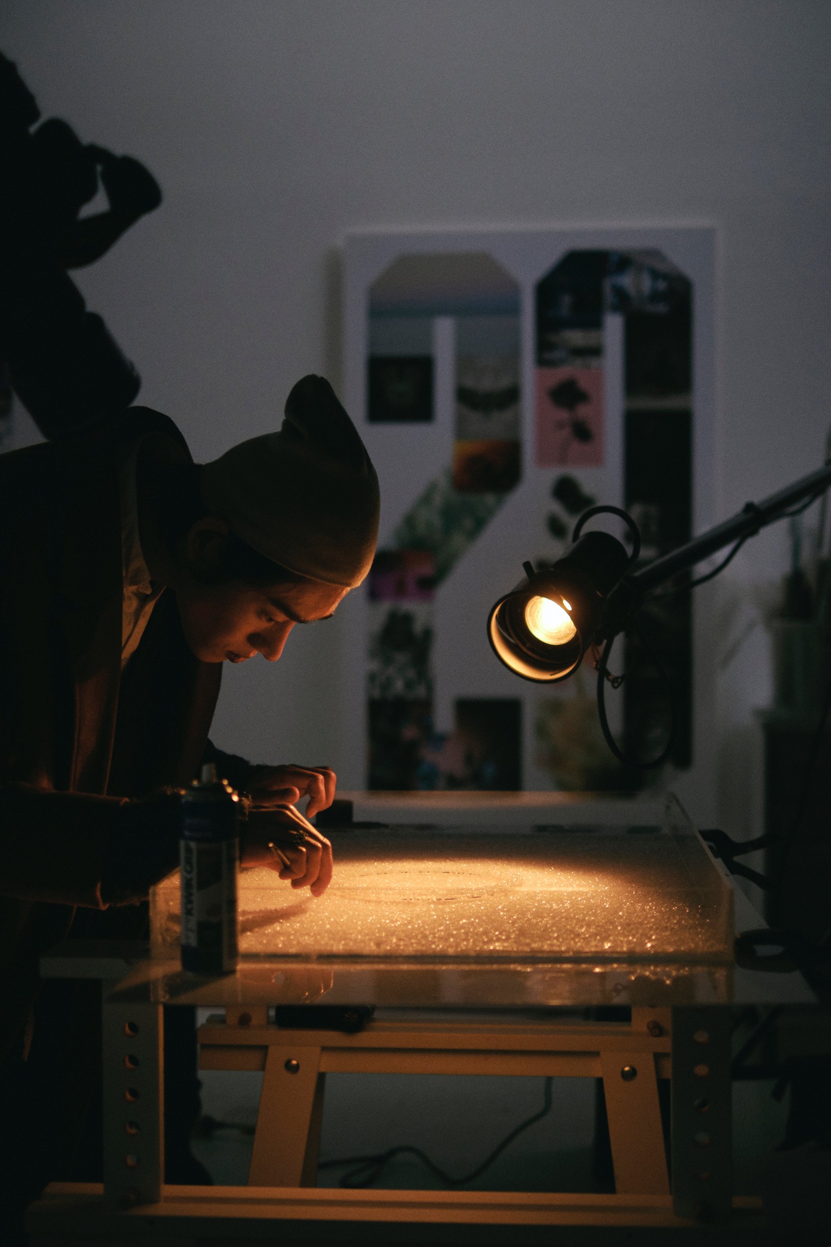 A person working on an art project in a dimly lit room, illuminated by a focused desk lamp, with a collage or photo collage in the background.