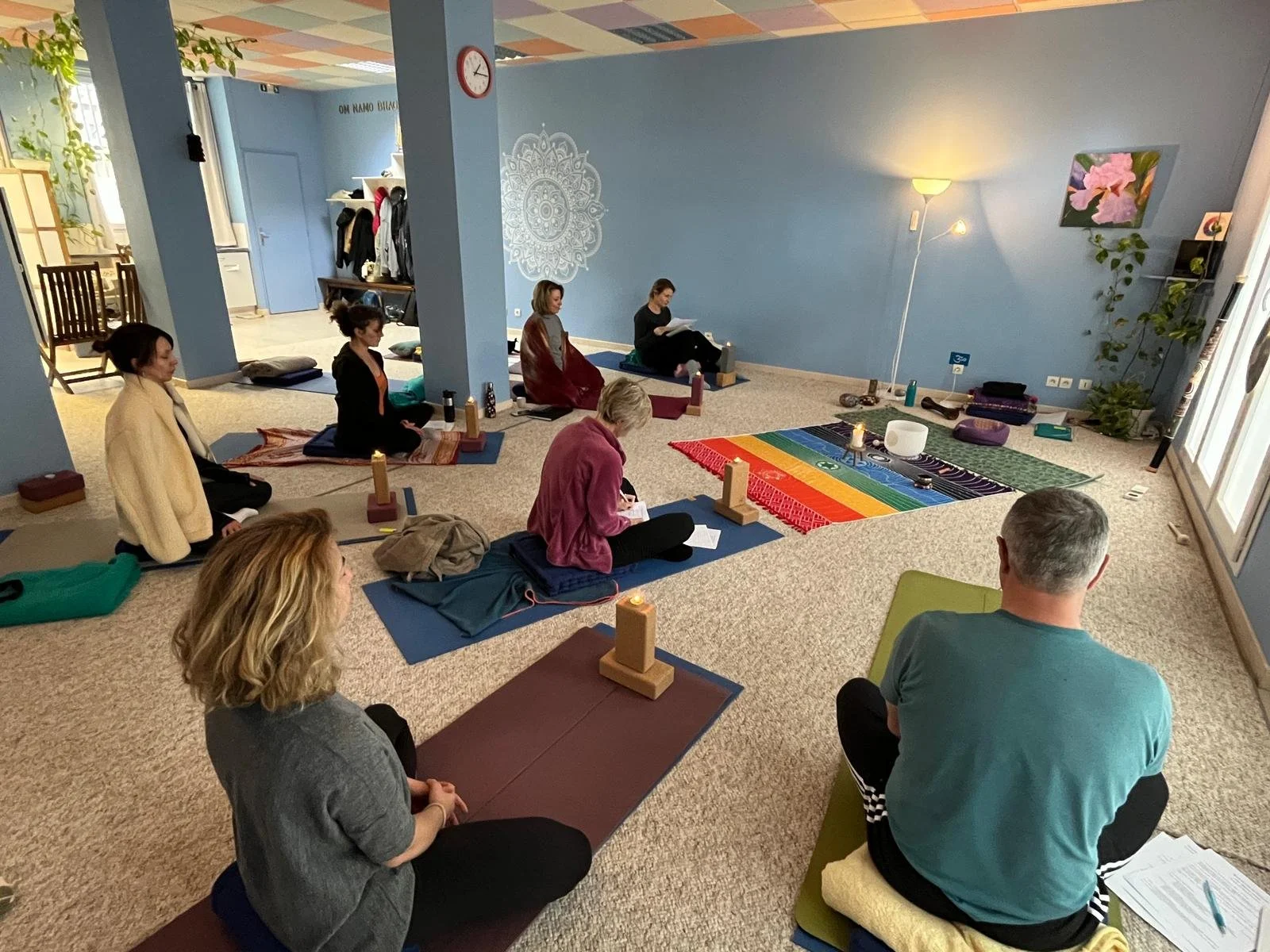 Groupe de personnes participant à une séance de méditation ou de yoga dans une pièce calme, assises sur des tapis, avec des bougies et des objets de spiritualité autour, dans une ambiance relaxante.