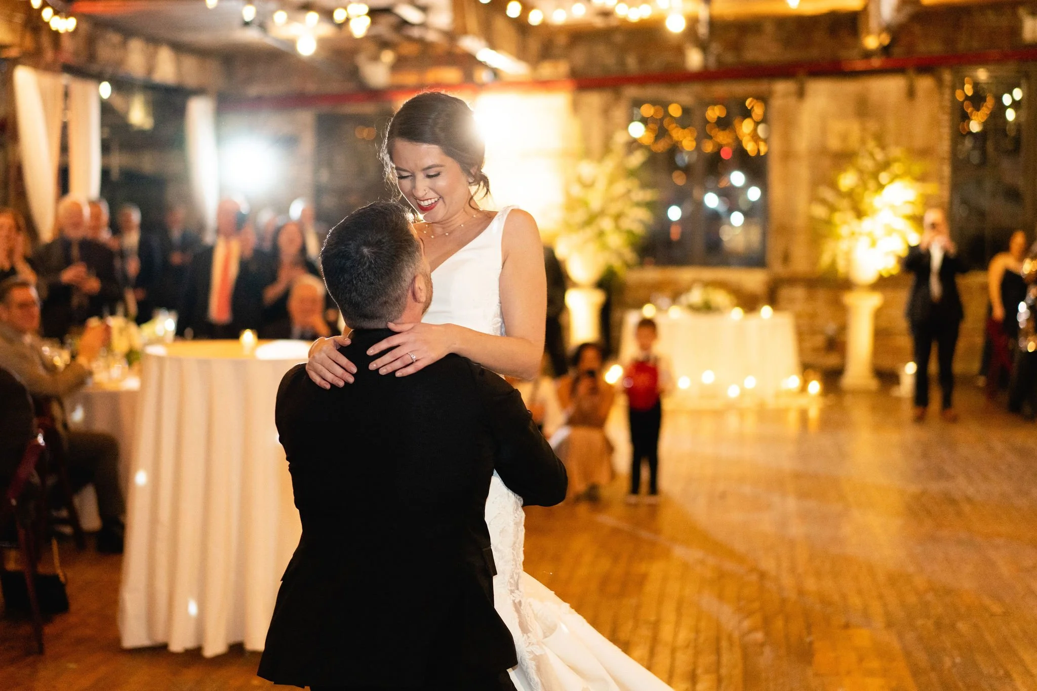 Couple dancing at a wedding reception with guests watching in a warmly lit, rustic venue.