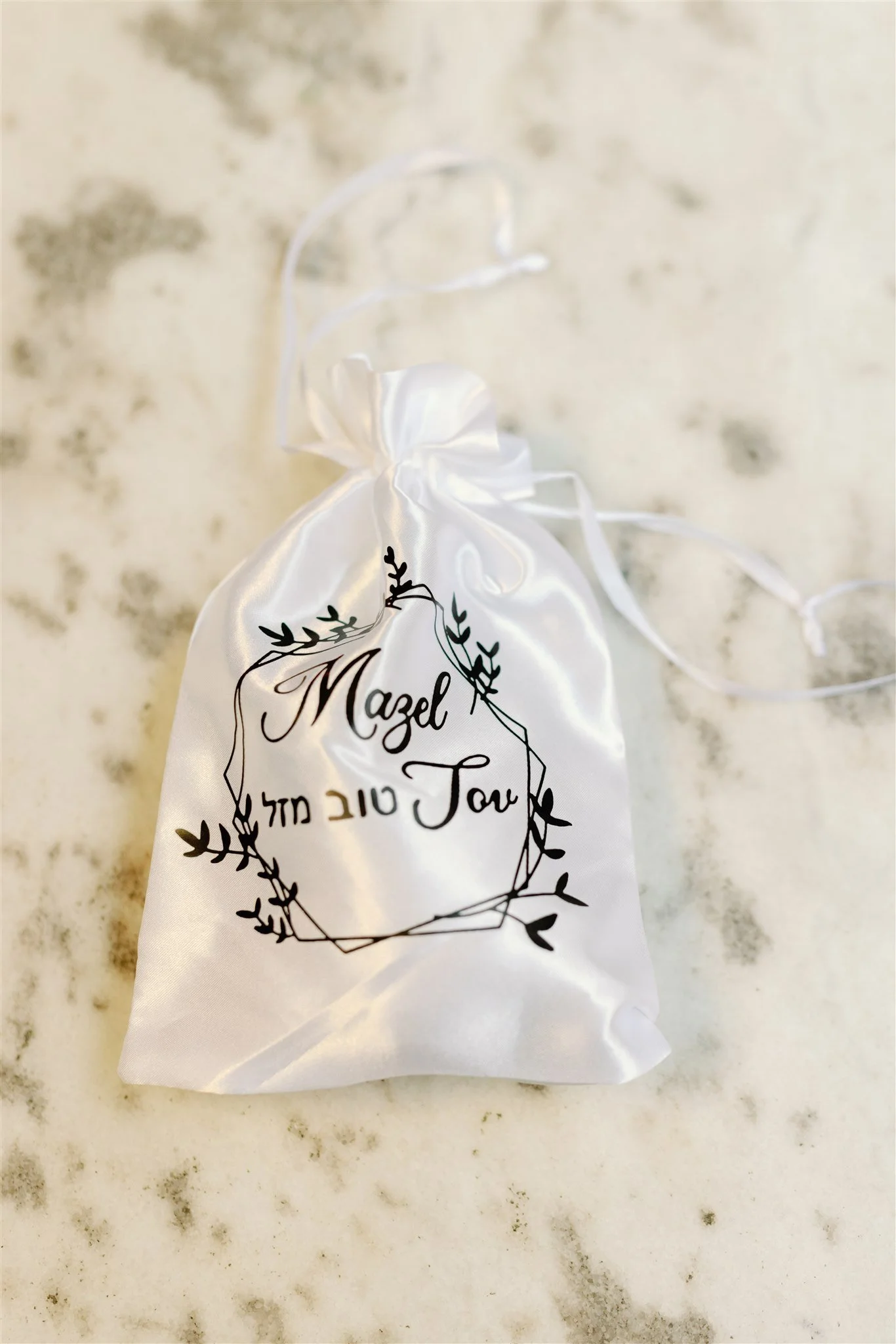 A white satin drawstring bag with black decorative writing and a botanical motif. The bag has the words 'Mazel Tov' and 'טוב לב' printed on it, indicating a celebratory occasion.