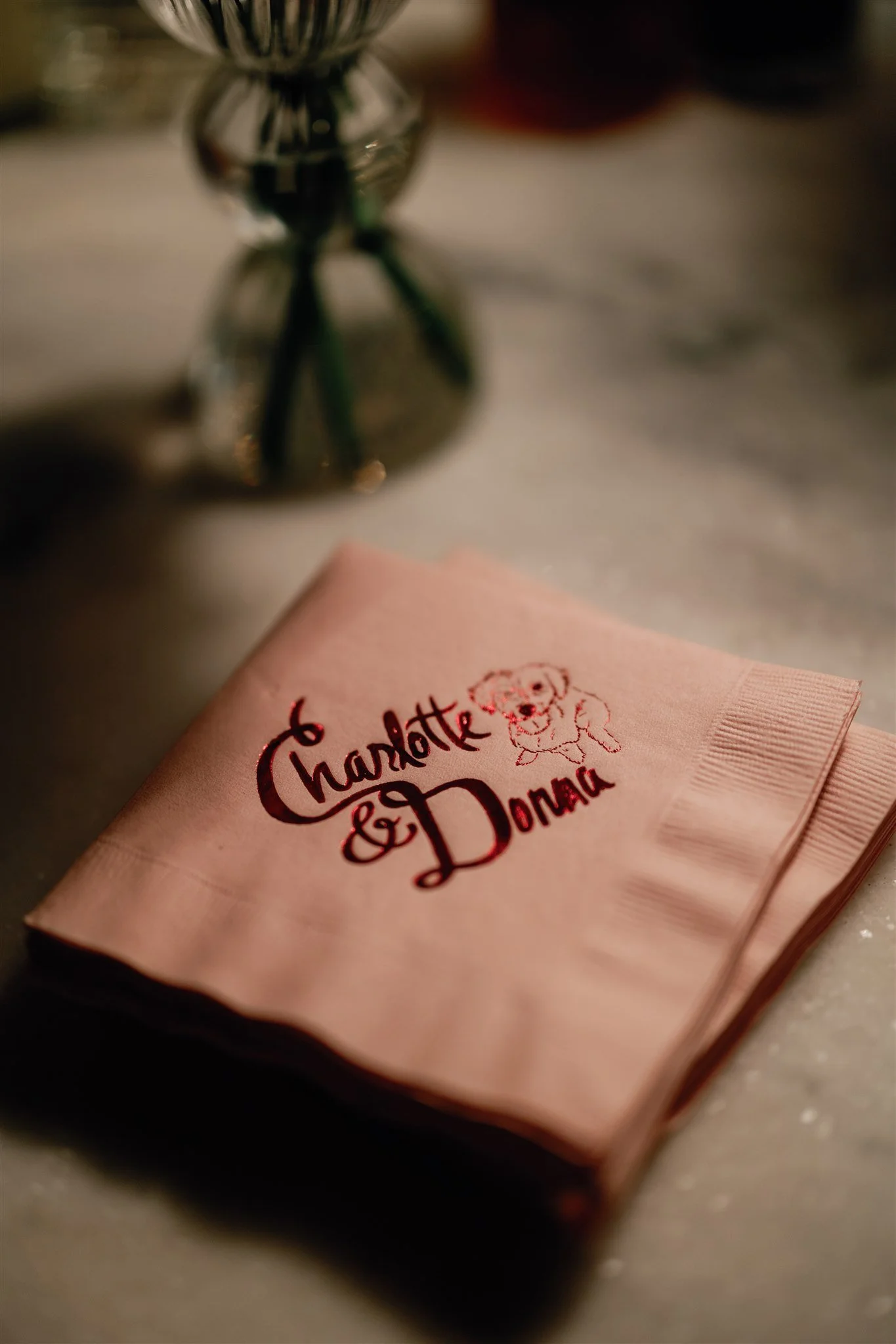 A pink napkin with a drawing of a dog and the words "Charlotte & Donna" written on it, resting on a surface with a blurred glass object in the background.