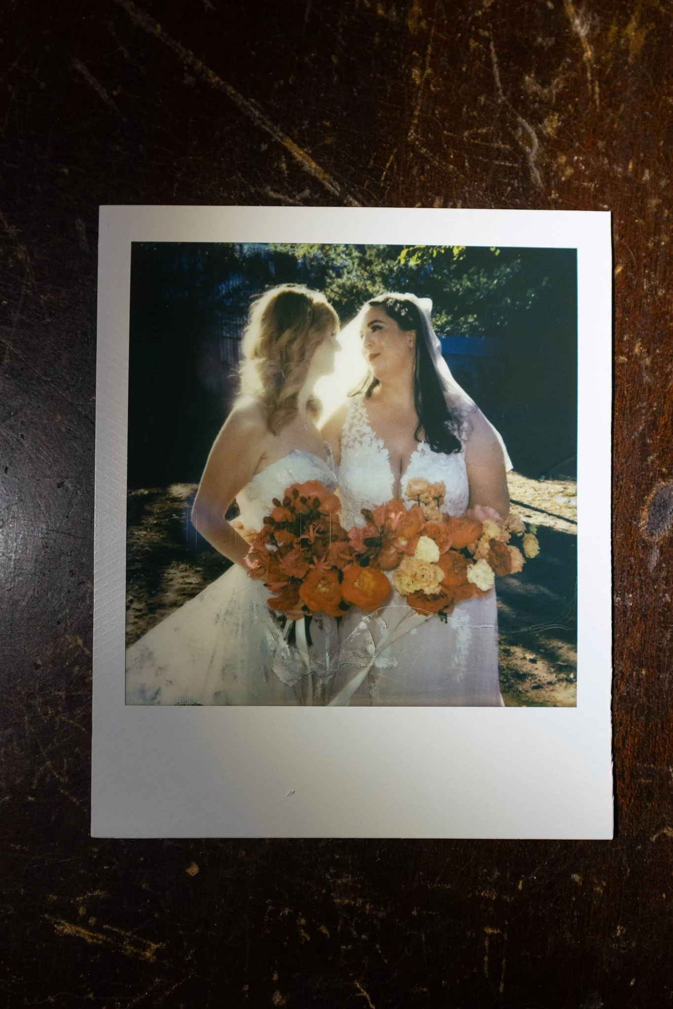 Two women in wedding dresses smiling and holding bouquets of orange, yellow, and red flowers, standing outdoors with sunlight filtering through trees in the background.