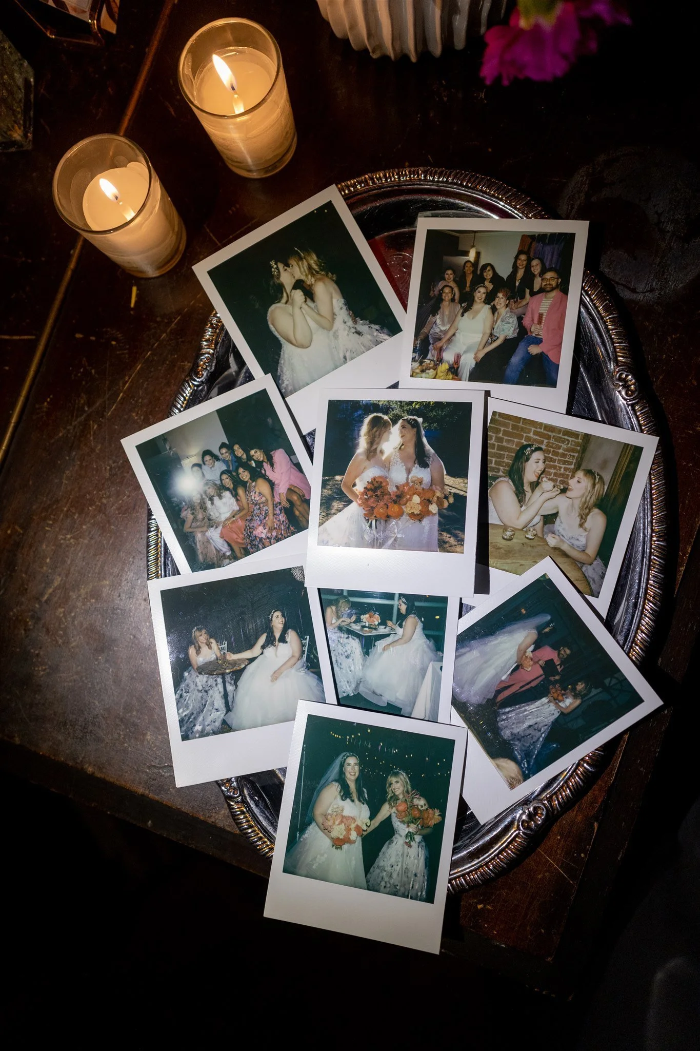 A collection of printed wedding photos spread on a dark wood table with two lit candles and which appears to be a silver tray underneath.