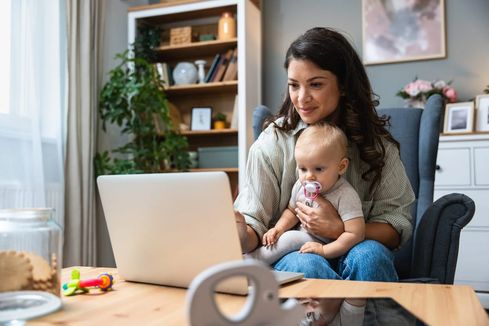 A woman and a baby sitting together in a living room, looking at a laptop screen. The woman is holding the baby on her lap, and the baby has a pacifier in their mouth. There are some toys on the table, and the background shows a bookshelf, plants, and framed pictures.