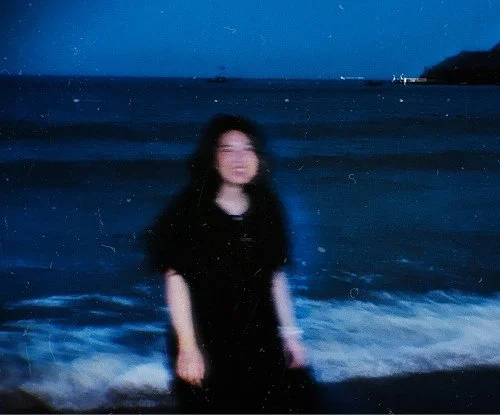 A person standing at the beach during nighttime with the ocean and sailboats in the background.