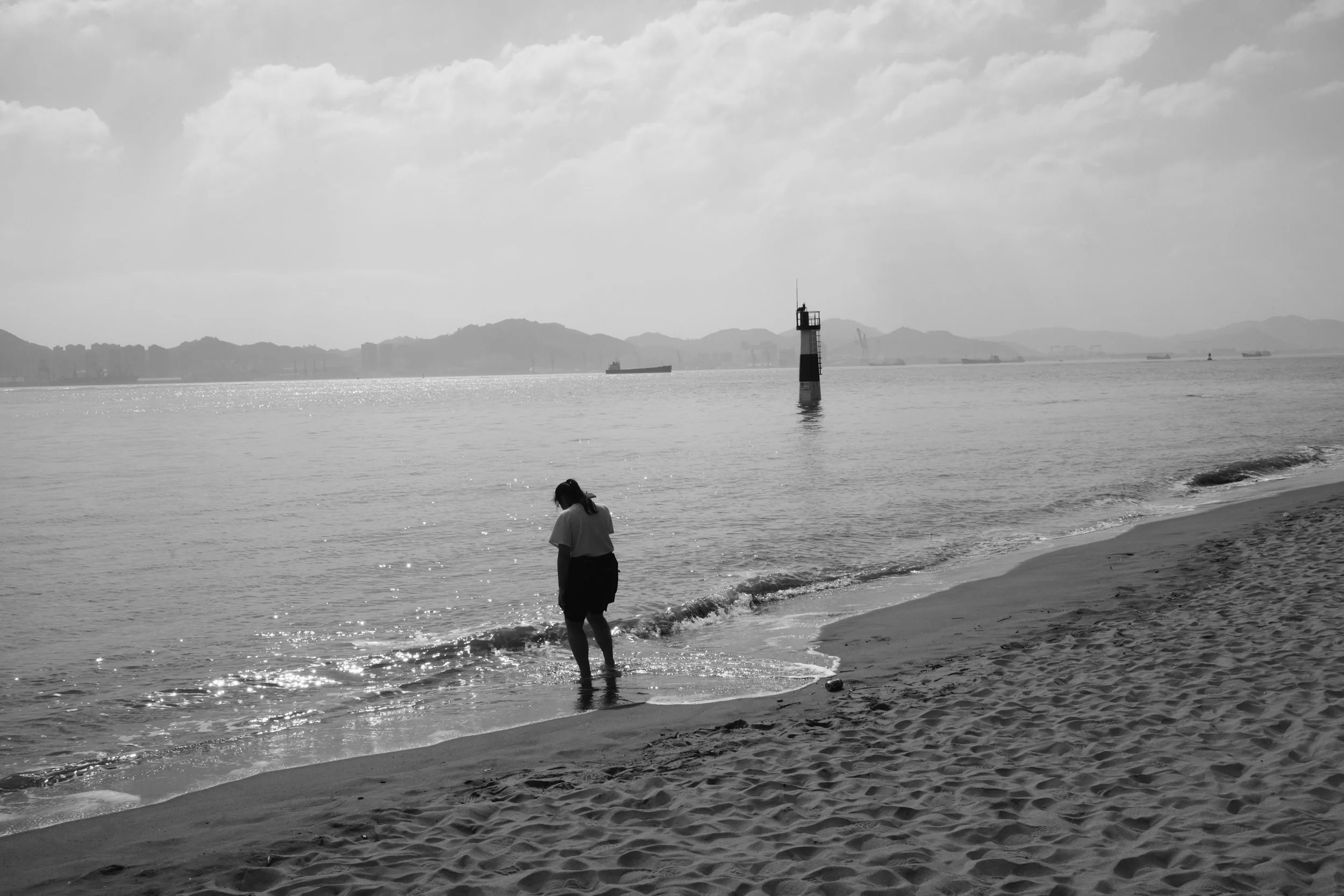 A person walking in shallow water at the beach with a lighthouse in the distance, under a cloudy sky, in black and white.