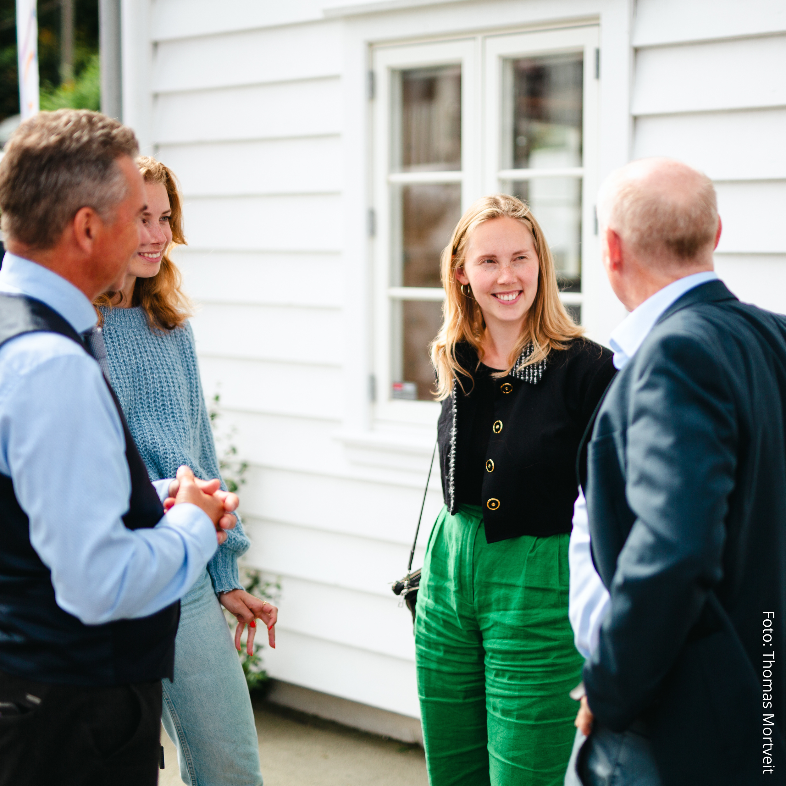 Fire personer som smiler til hverandre utenfor et hvitt hus. Fremje.