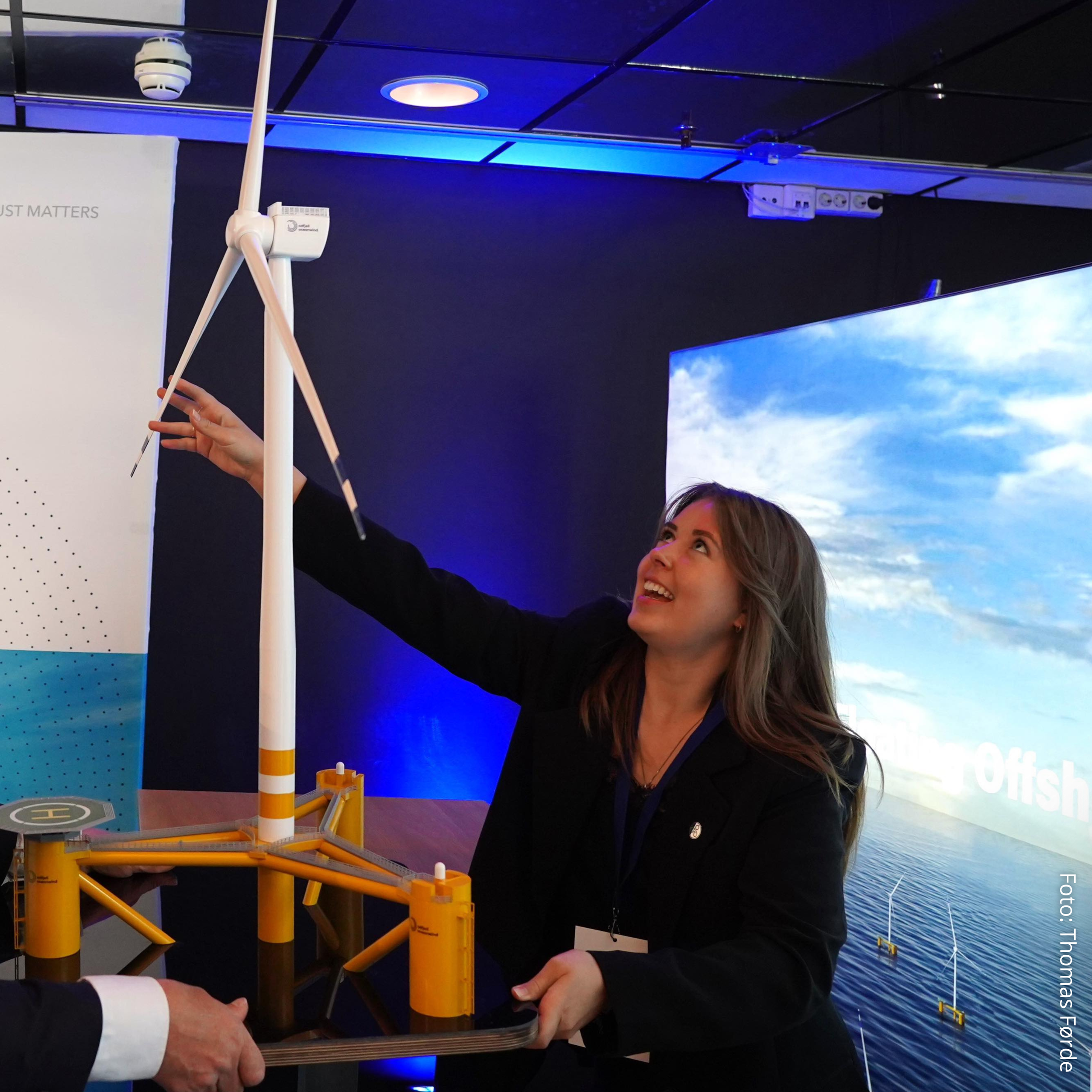 En kvinne smiler mens hun peker på en modell av en vindmølle. Bakgrunn med bilde av hav og himmel.