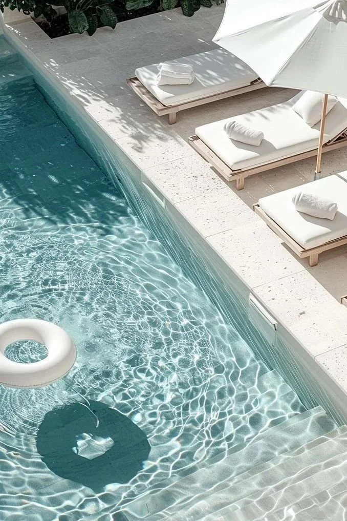 Piscine avec transats et parasol blanc, serviettes pliées, et reflet de lumière sur l'eau.