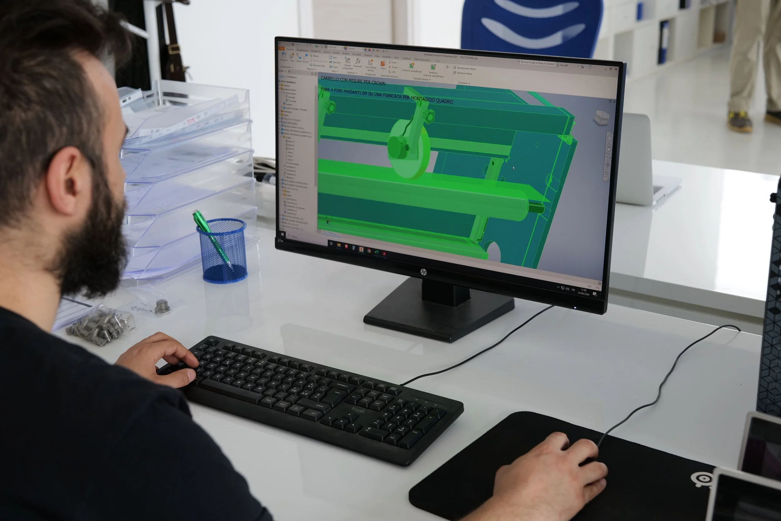 A man with a beard working at a computer in an office. The screen displays a 3D model of a mechanical component in green and blue colors. On the desk are small metal parts, a blue pen in a mesh pen holder, and clear plastic trays.