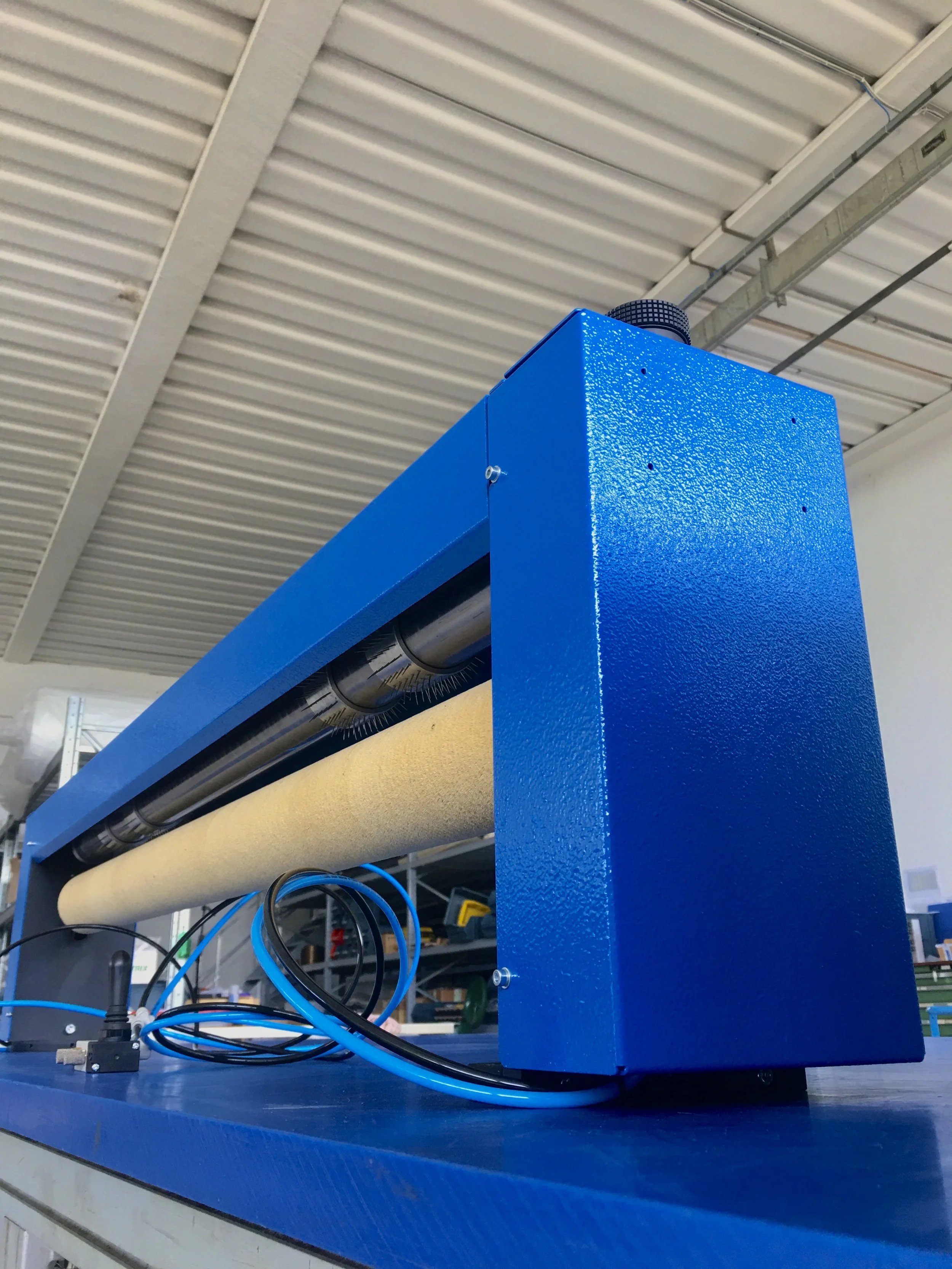 Blue industrial machine or component with a roller, wires, and a textured surface, located in a warehouse or workshop with a metal ceiling.